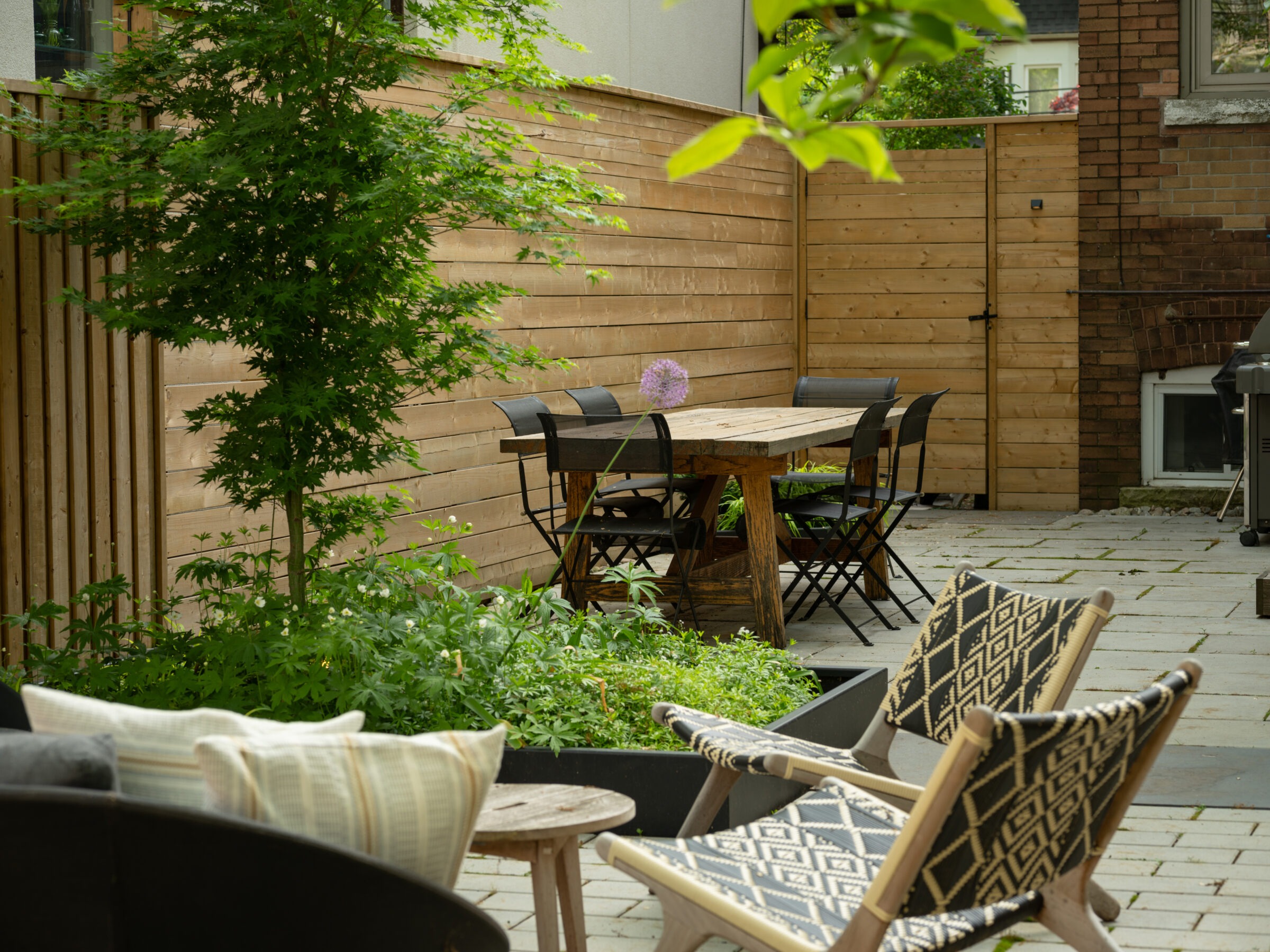 Cozy outdoor patio with wooden table, chairs, and planters. Relaxing arranged seating with patterned chairs. Lush greenery and a wooden fence.