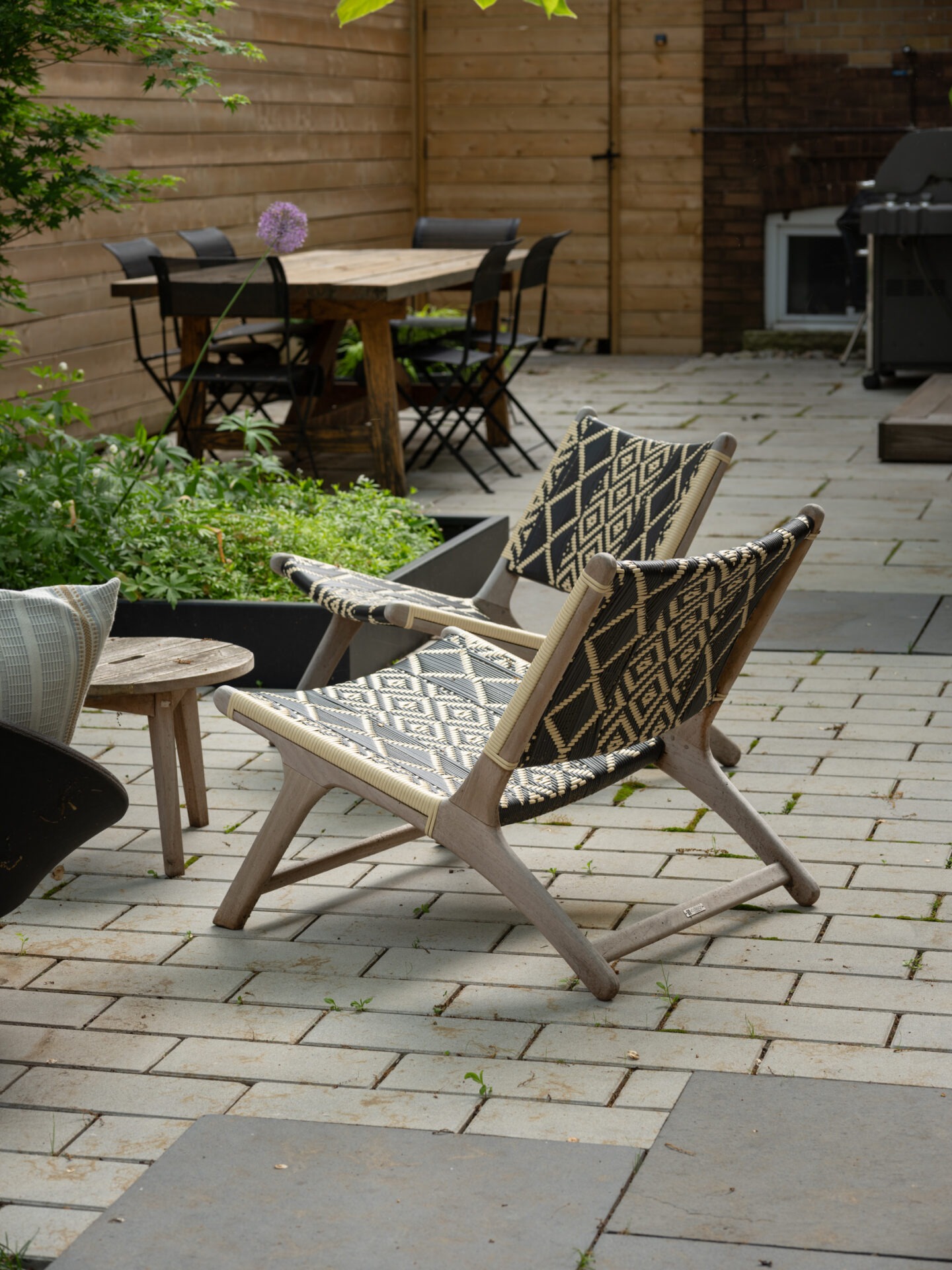 Cozy patio with patterned chairs, wooden table, and lush greenery. Brick and wooden walls create a serene, inviting outdoor space for relaxation.