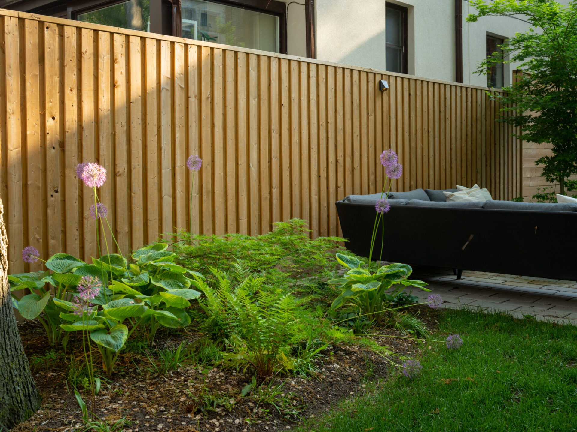 A cozy garden with a wooden fence, purple flowers, lush greenery, and a modern outdoor sofa, creates a serene ambiance.