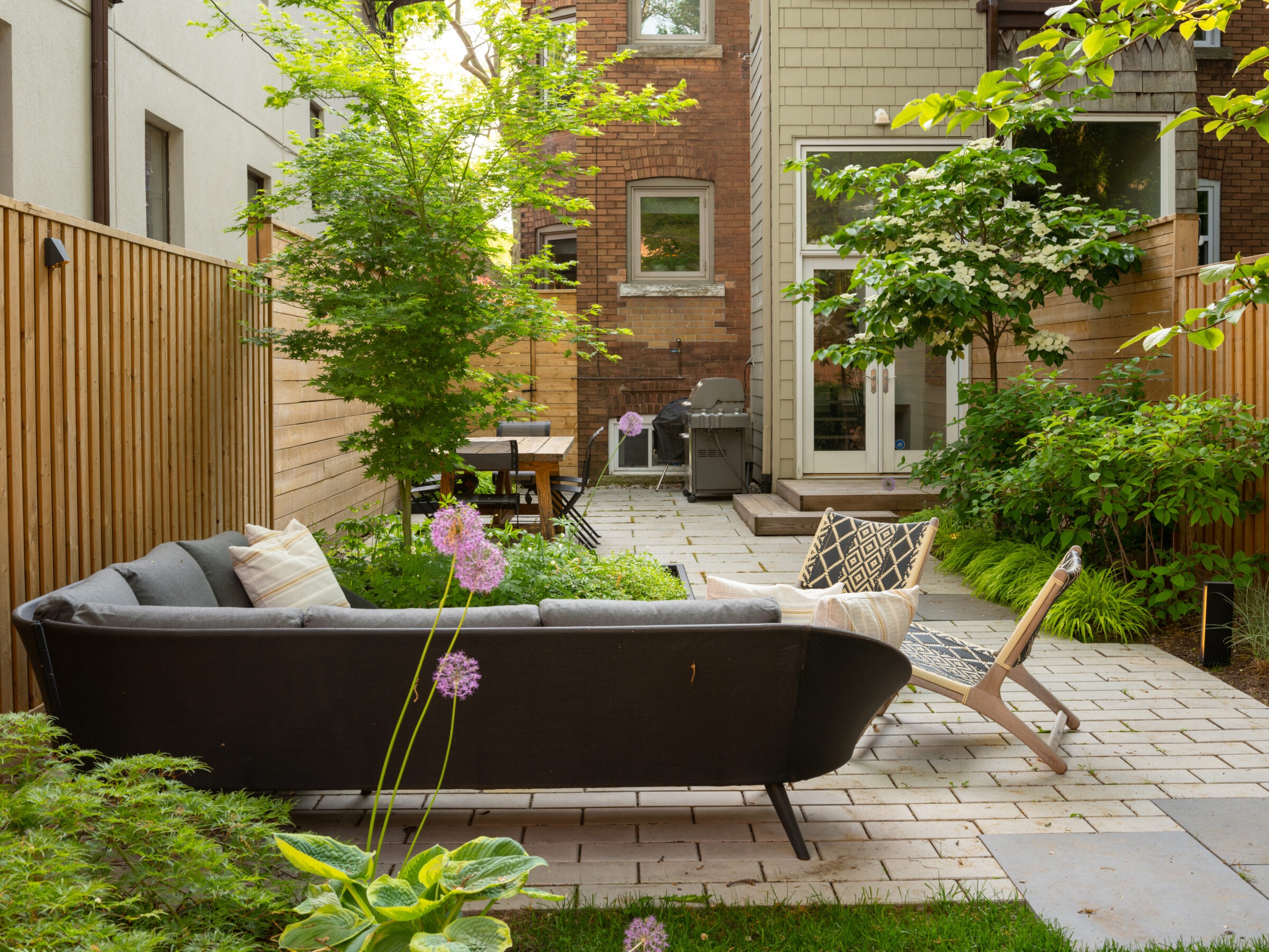Cozy backyard patio with seating, surrounded by lush greenery and wooden fencing. Brick and wood building exterior in the background. Peaceful atmosphere.