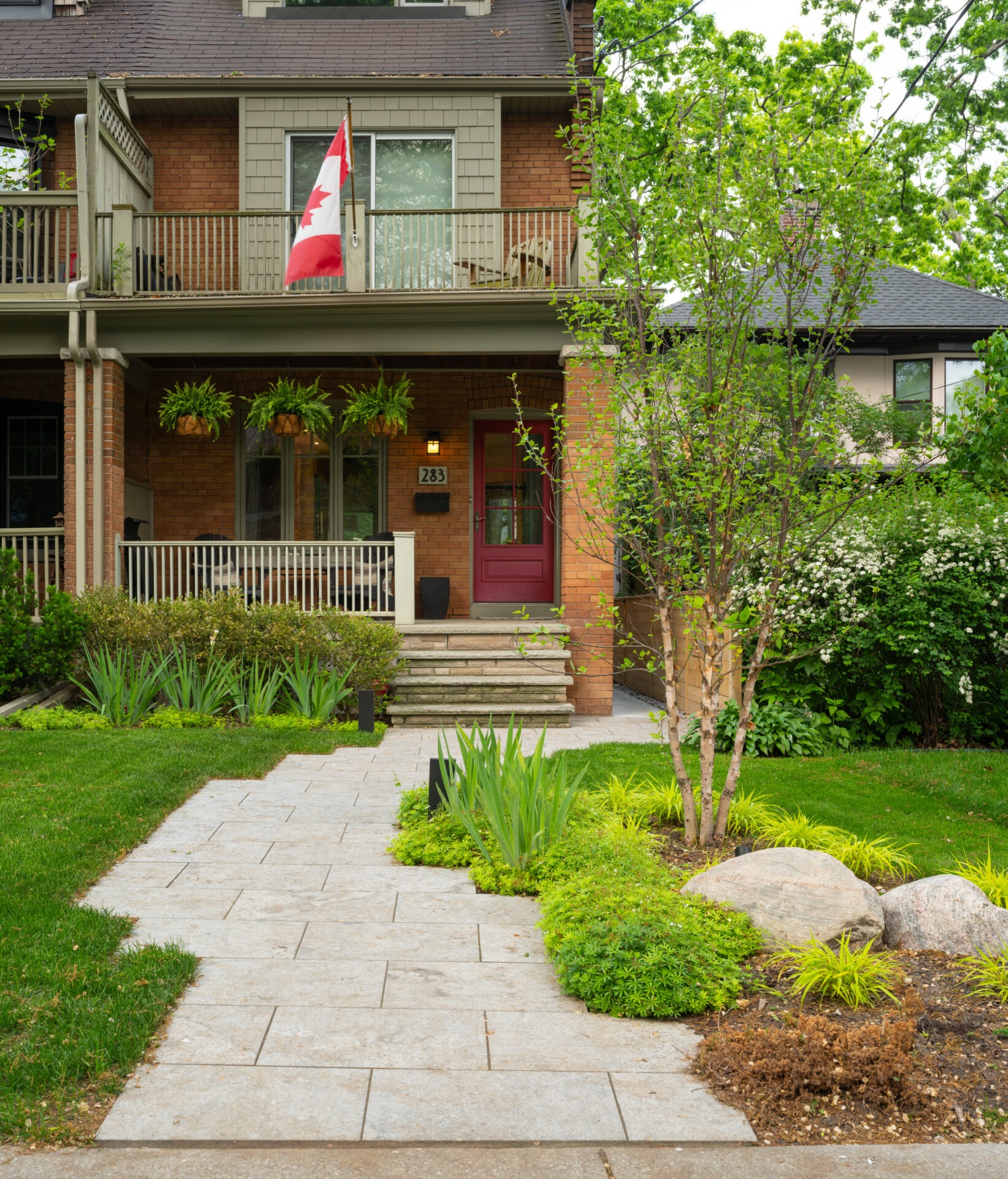 A charming brick house with a Canadian flag, porch seating, landscaped garden, and stone pathway, surrounded by lush greenery and trees.