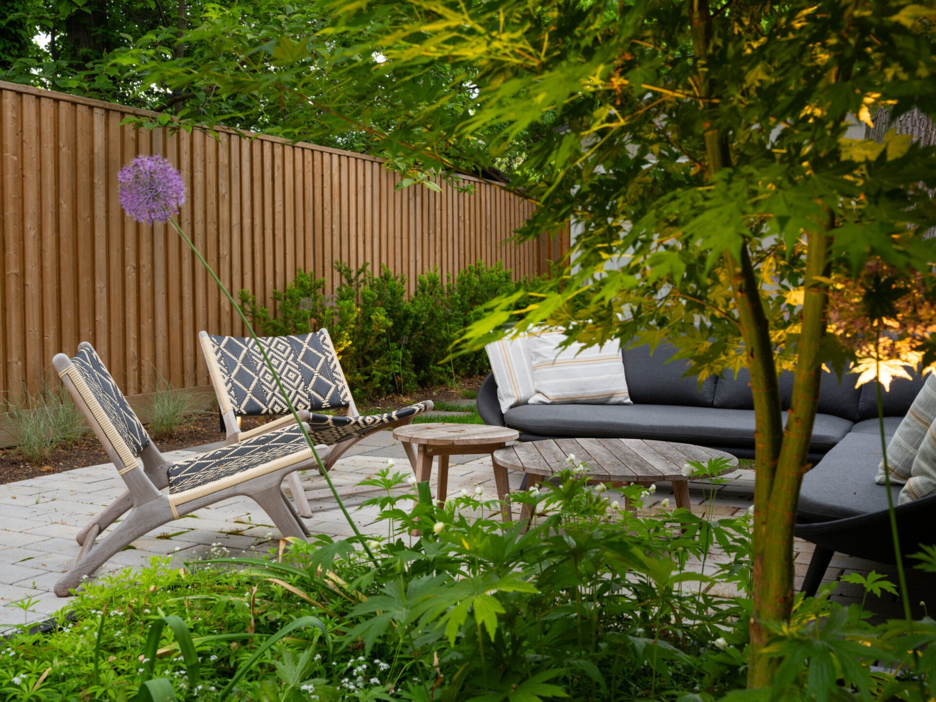 A cozy outdoor patio with patterned chairs, wooden table, lush greenery, and a wooden fence, illuminated by warm lighting at dusk.