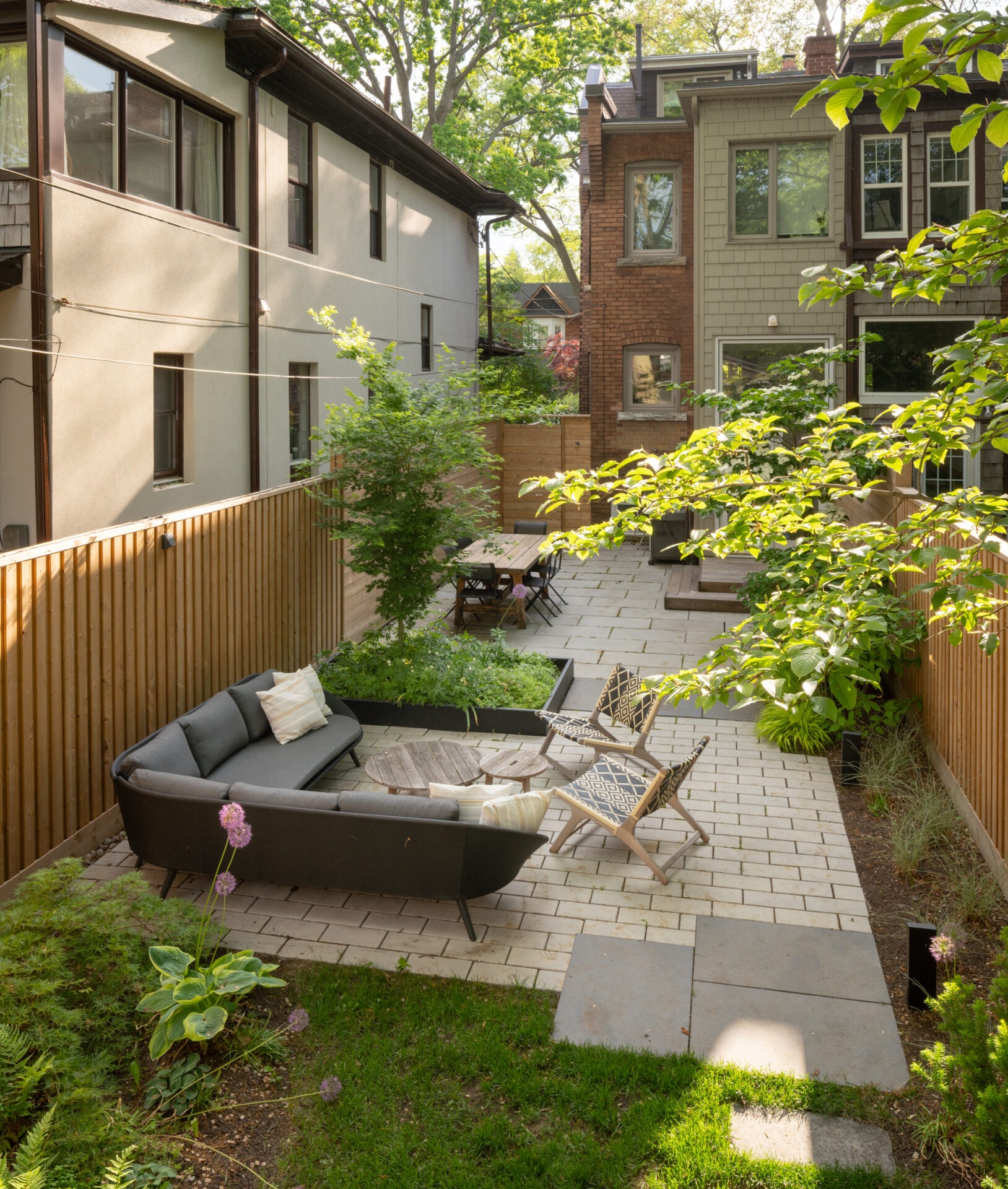 A backyard patio with modern furniture, lush greenery, and potted plants. Wooden fence surrounds the area, with a table and chairs in view.