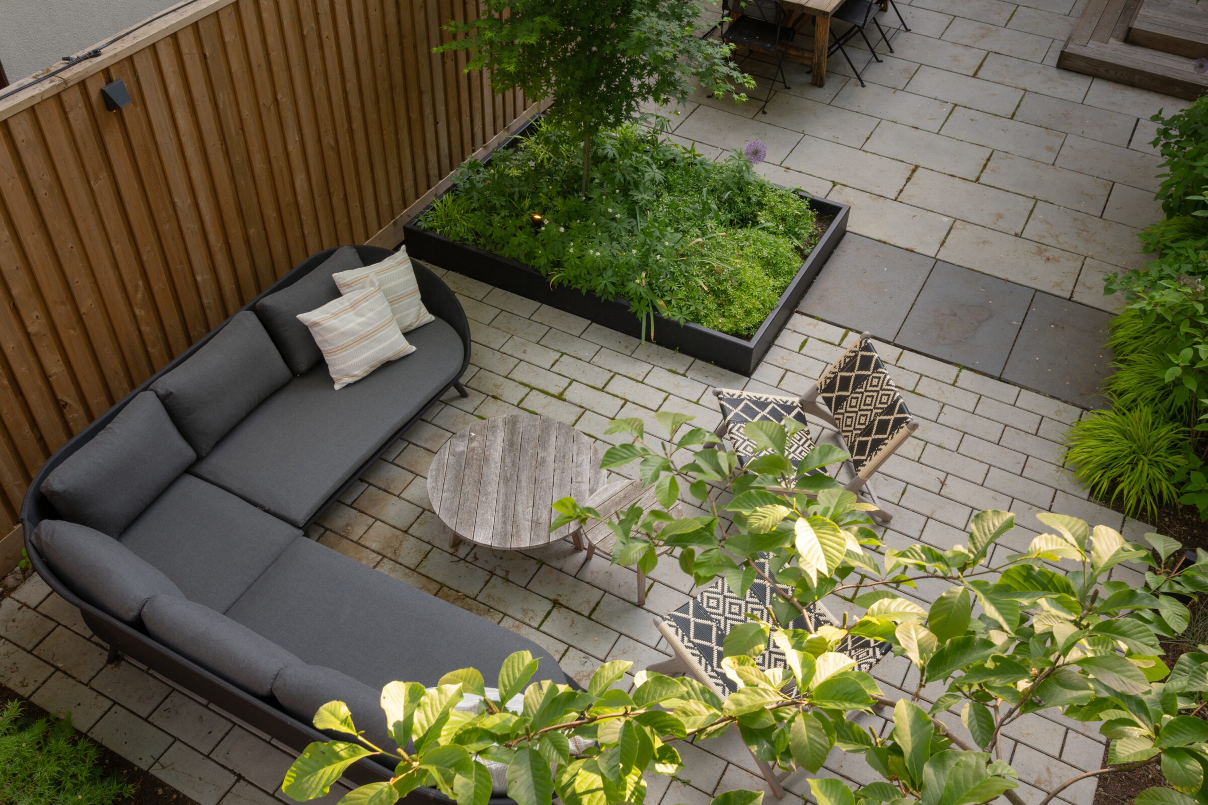 A cozy patio features a large gray couch, wooden round table, patterned chairs, and plants. Wooden fencing and pavers complete the serene setting.