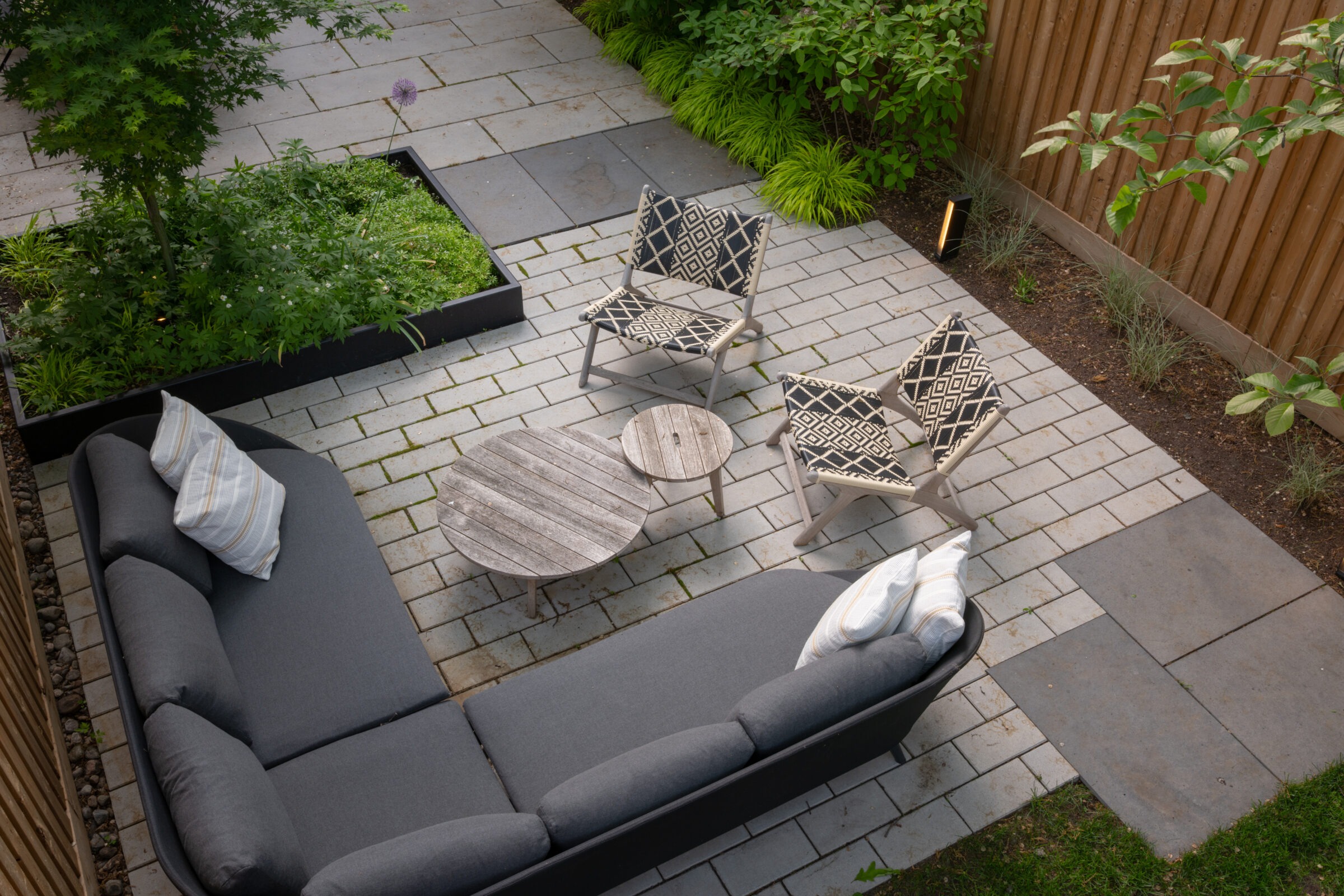 Modern garden patio with gray sectional sofa, patterned chairs, wooden tables, and lush greenery. Stylish outdoor gathering space with comfortable seating.