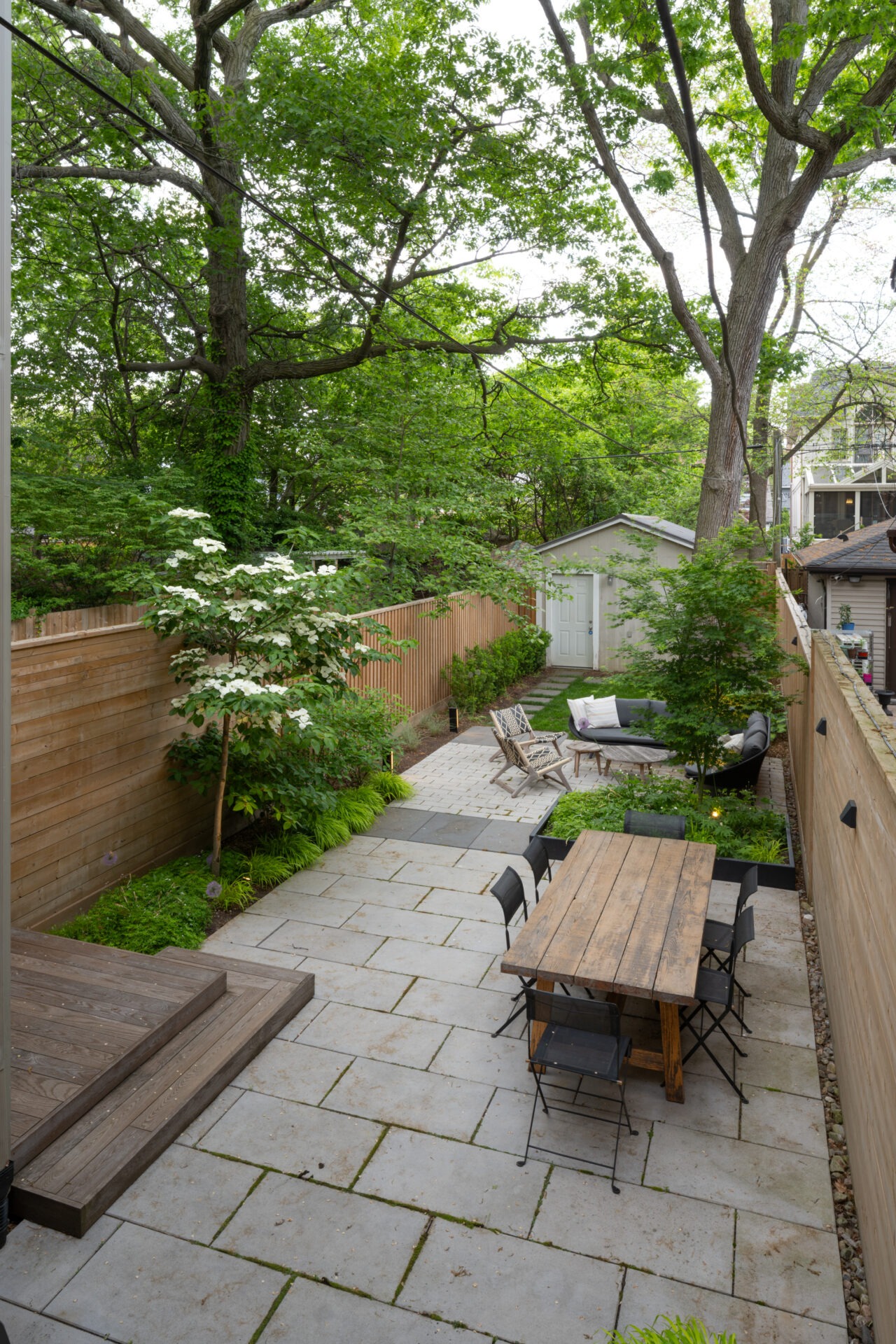 A serene backyard features a patio with wooden dining table, chairs, garden, and surrounding trees, all enclosed by a wooden fence.