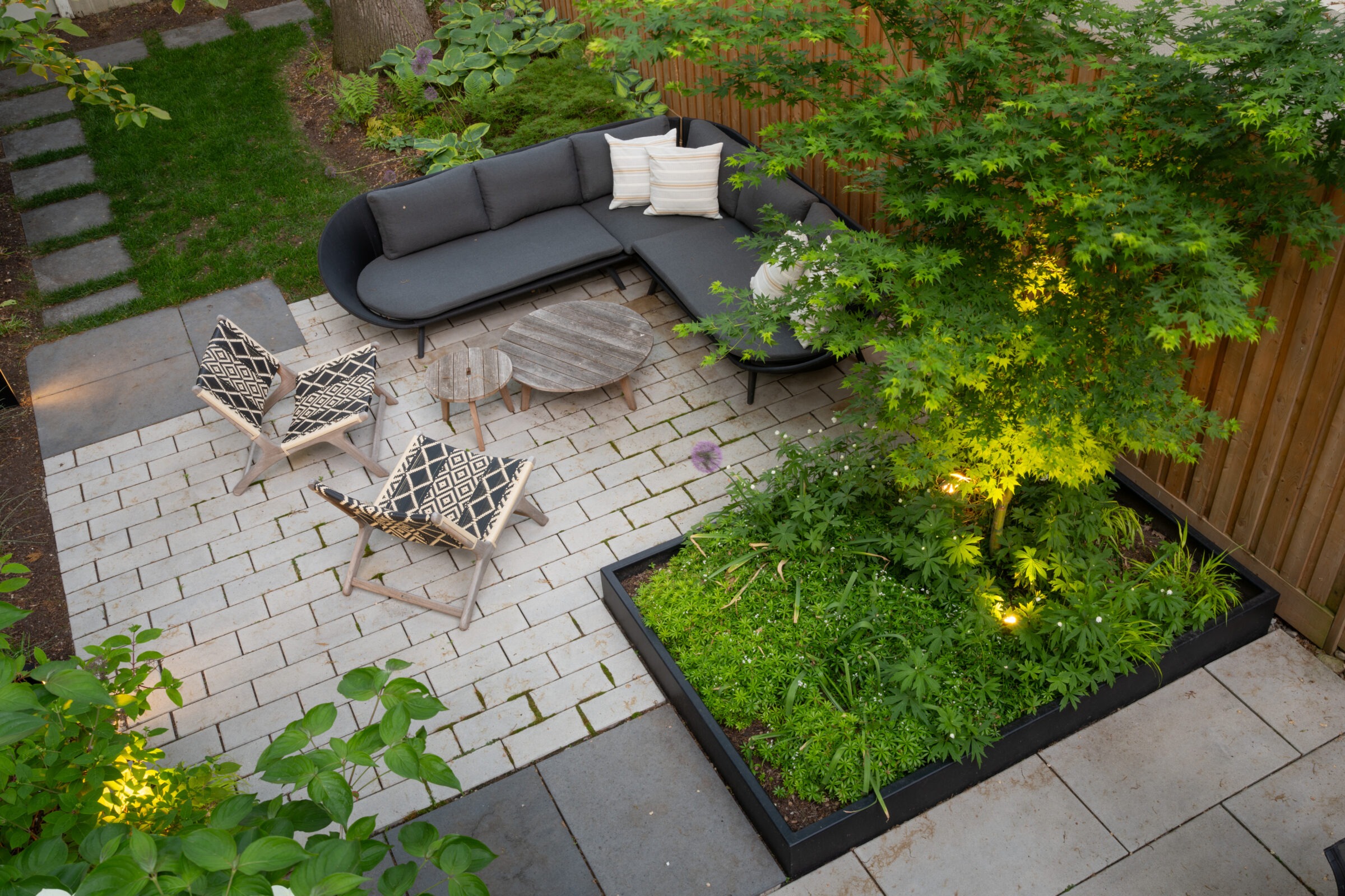 Cozy backyard with modern patio furniture, geometric-patterned chairs, and lush greenery. Soft ambient lighting highlights a tree and vibrant planter.