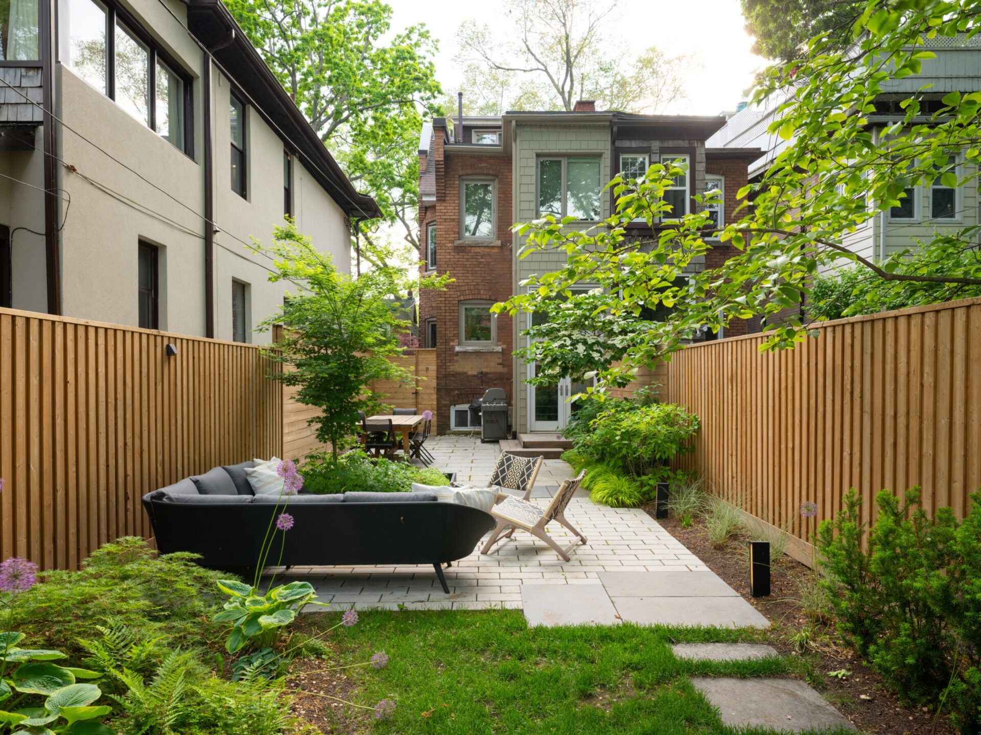 A cozy backyard features patio furniture, lush greenery, and wooden fencing, leading up to a charming brick house under a clear, sunny sky.