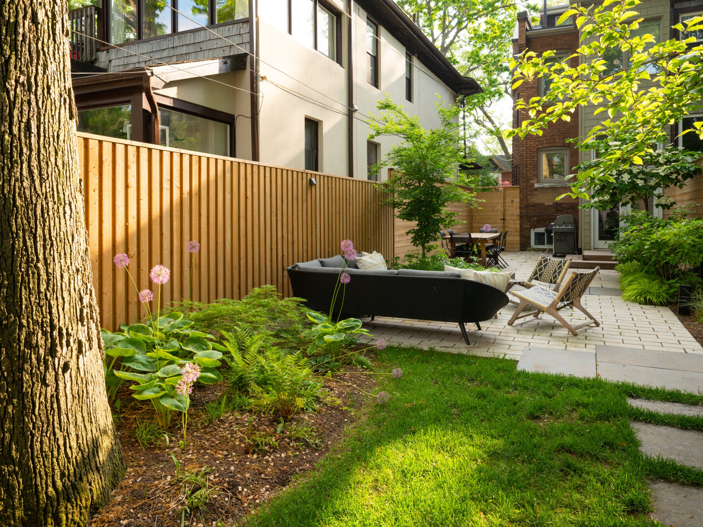 A cozy backyard with modern patio furniture, wooden fence, and lush greenery including ferns and flowering plants, creating a relaxing outdoor space.