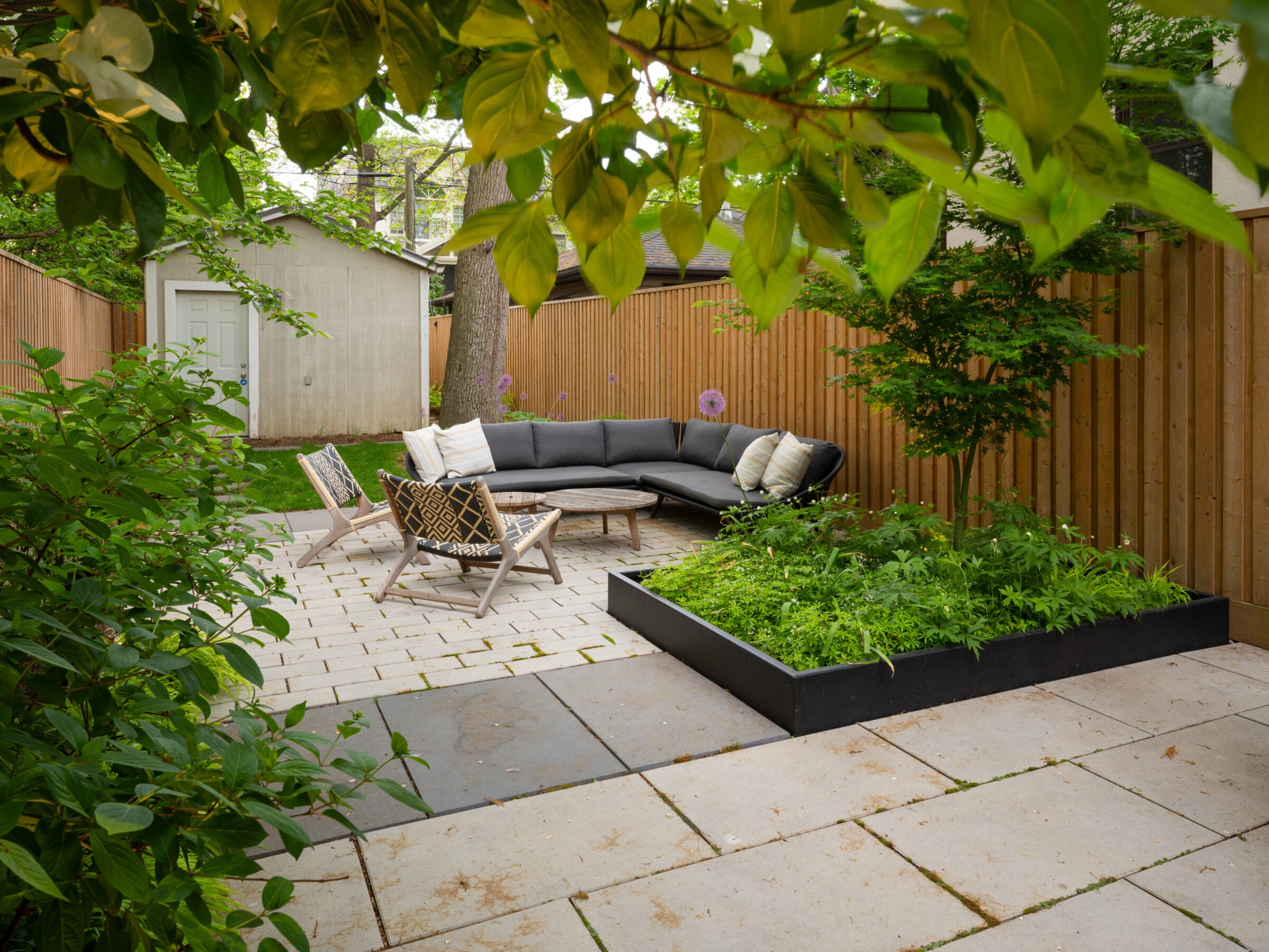 A peaceful, fenced backyard features a patio with modern seating, a small garden, and a shed, surrounded by lush greenery and trees.