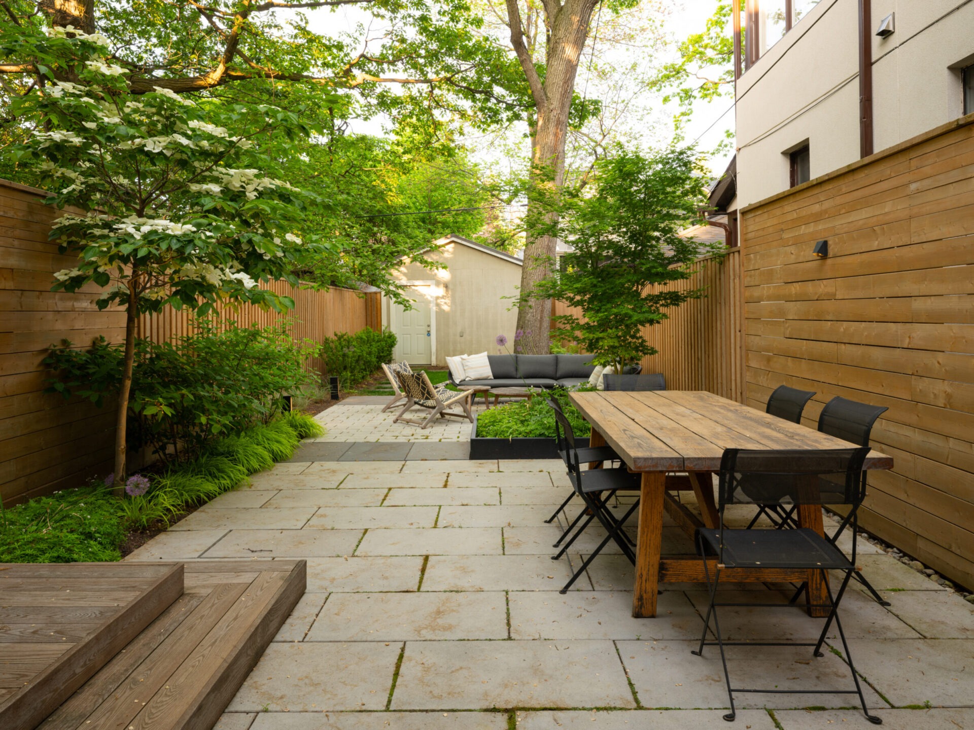 A tranquil backyard patio features a wooden table with chairs, surrounded by lush greenery and wooden fences, creating an inviting outdoor space.