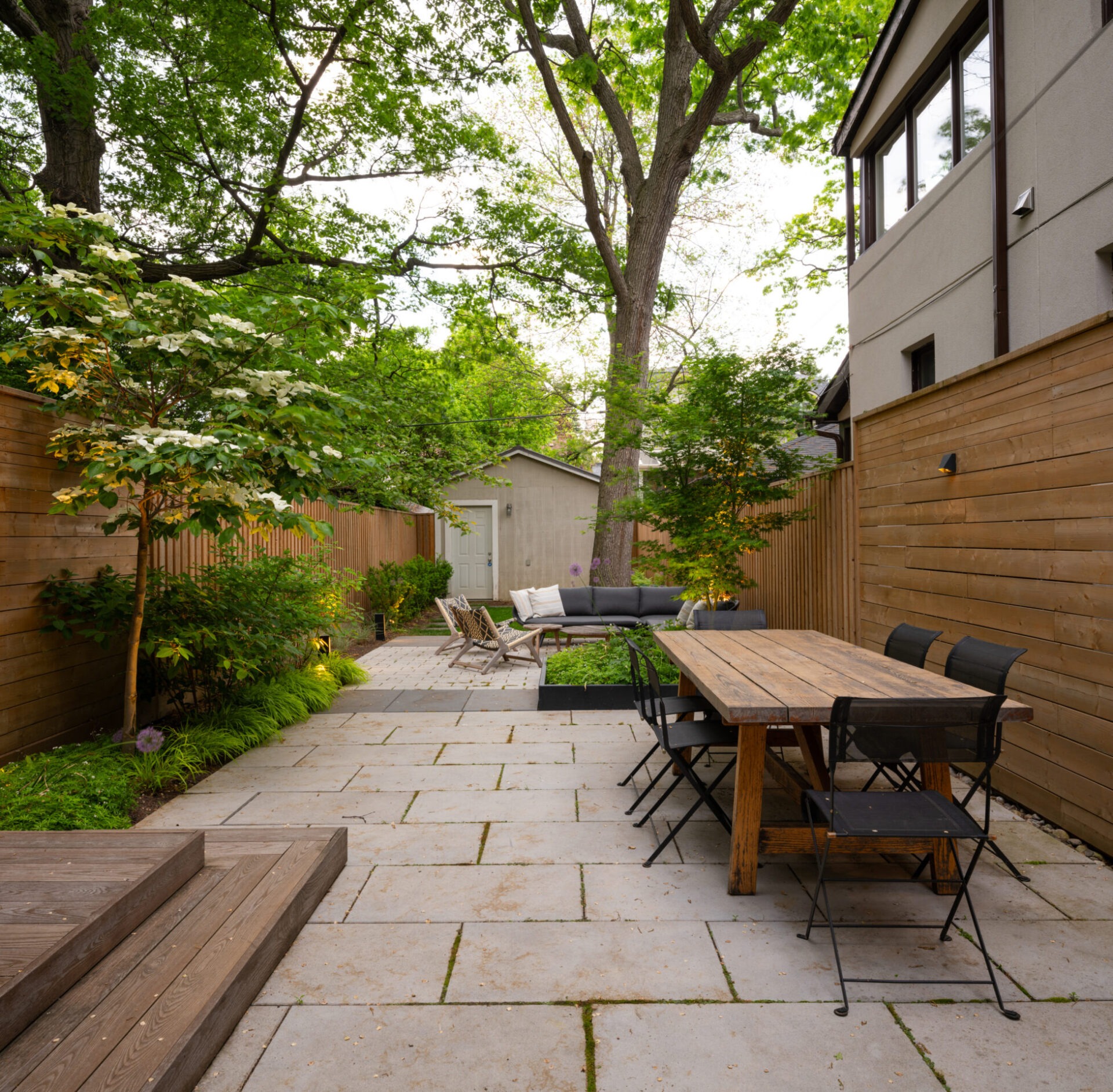Modern backyard with wooden dining table, chairs, cozy seating area, and lush greenery. Tall trees provide shade and natural beauty. Peaceful atmosphere.