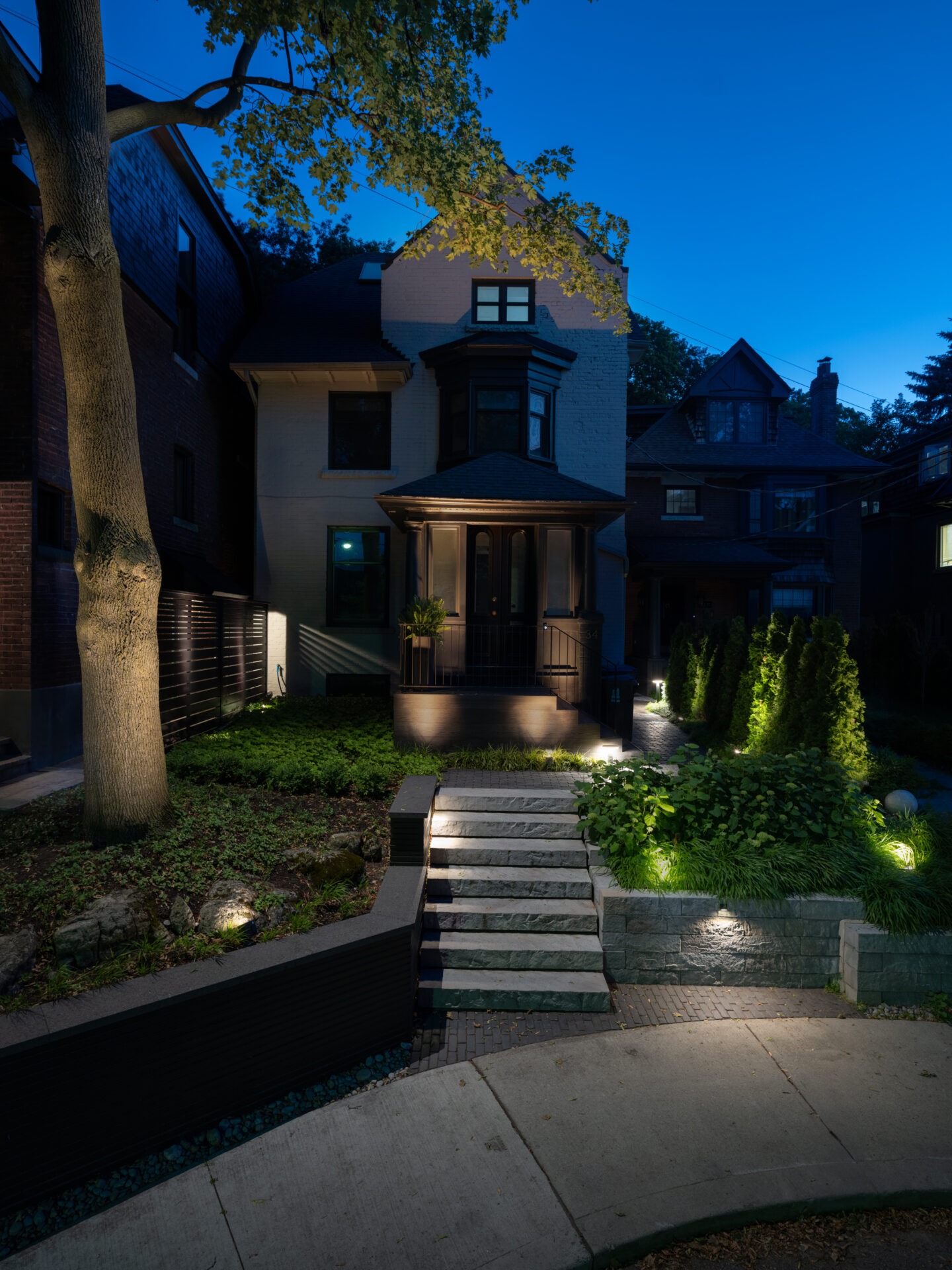 A charming house at dusk, featuring a lit pathway, garden, and mature trees; enveloped in evening tranquility, no landmarks visible.