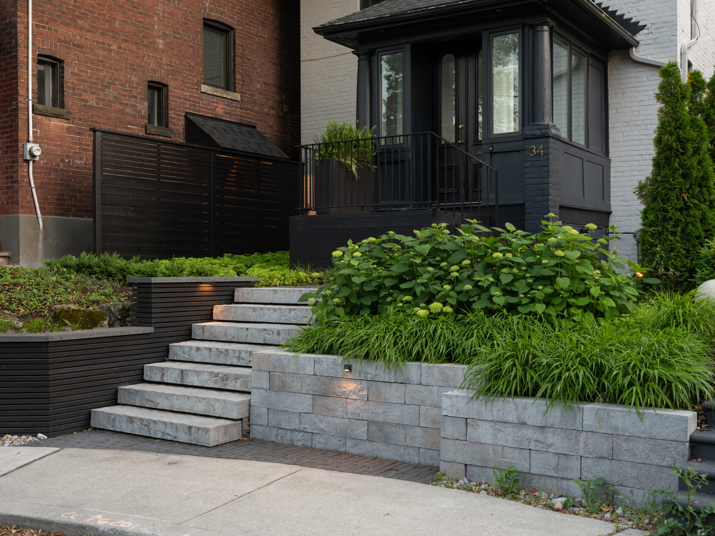 Stone steps leading to a porch surrounded by lush greenery, beside a brick building, with structured landscaping and ambient lighting.