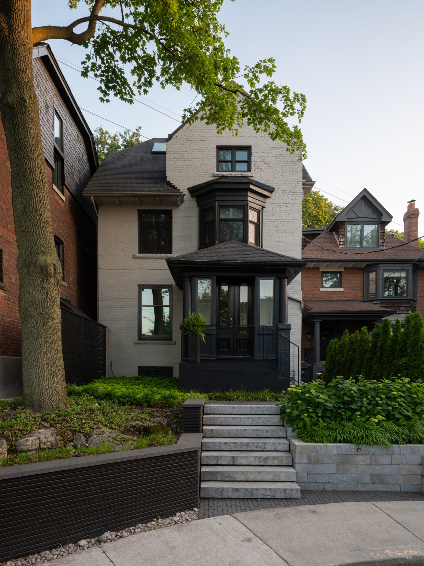 Victorian-style house with black accents, nestled between brick homes. Greenery surrounds the entrance, with steps leading up from the sidewalk.