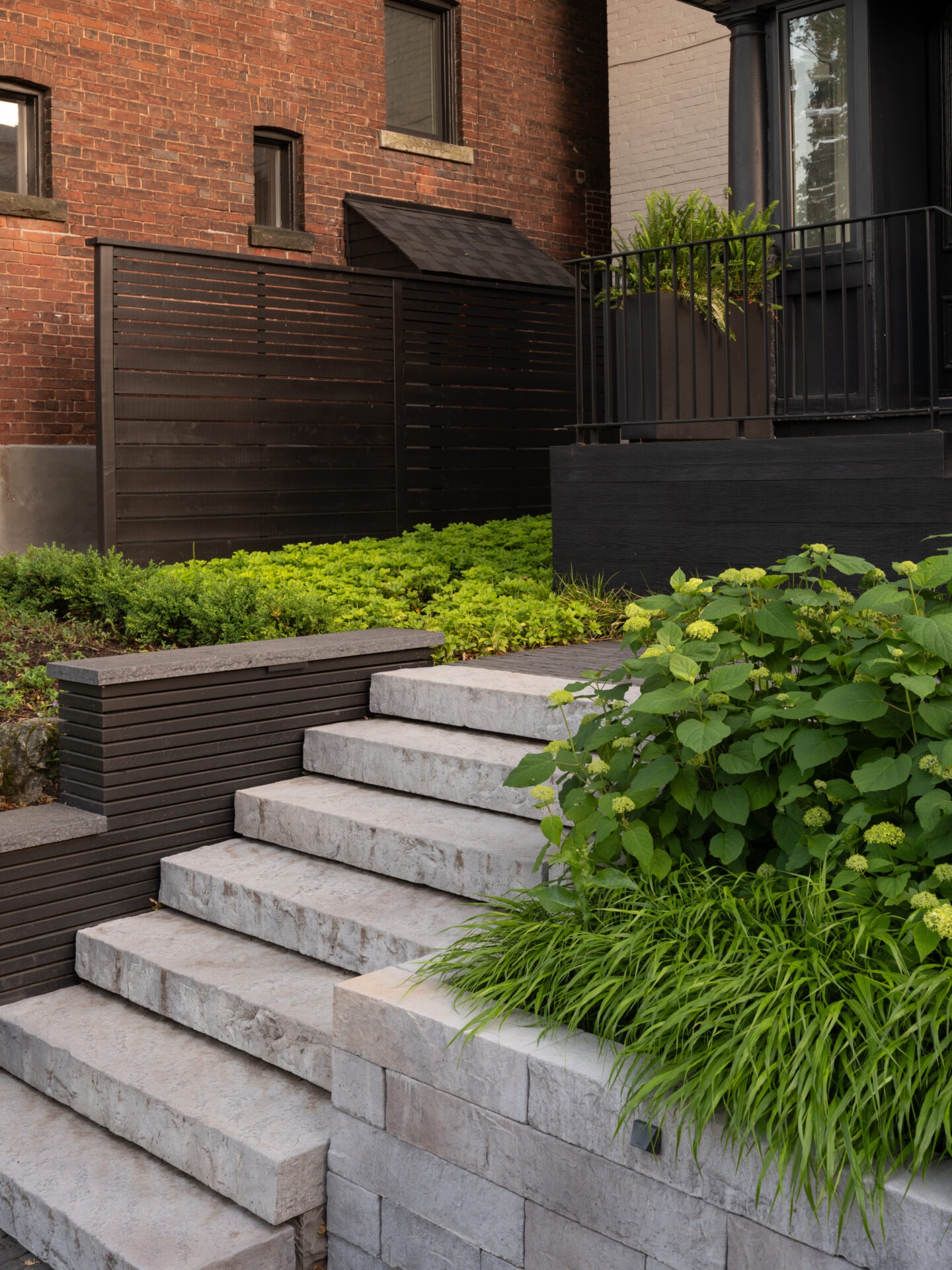 Brick townhouse with stone steps, black railing, and lush greenery. A wooden privacy screen separates the garden area from neighboring spaces.