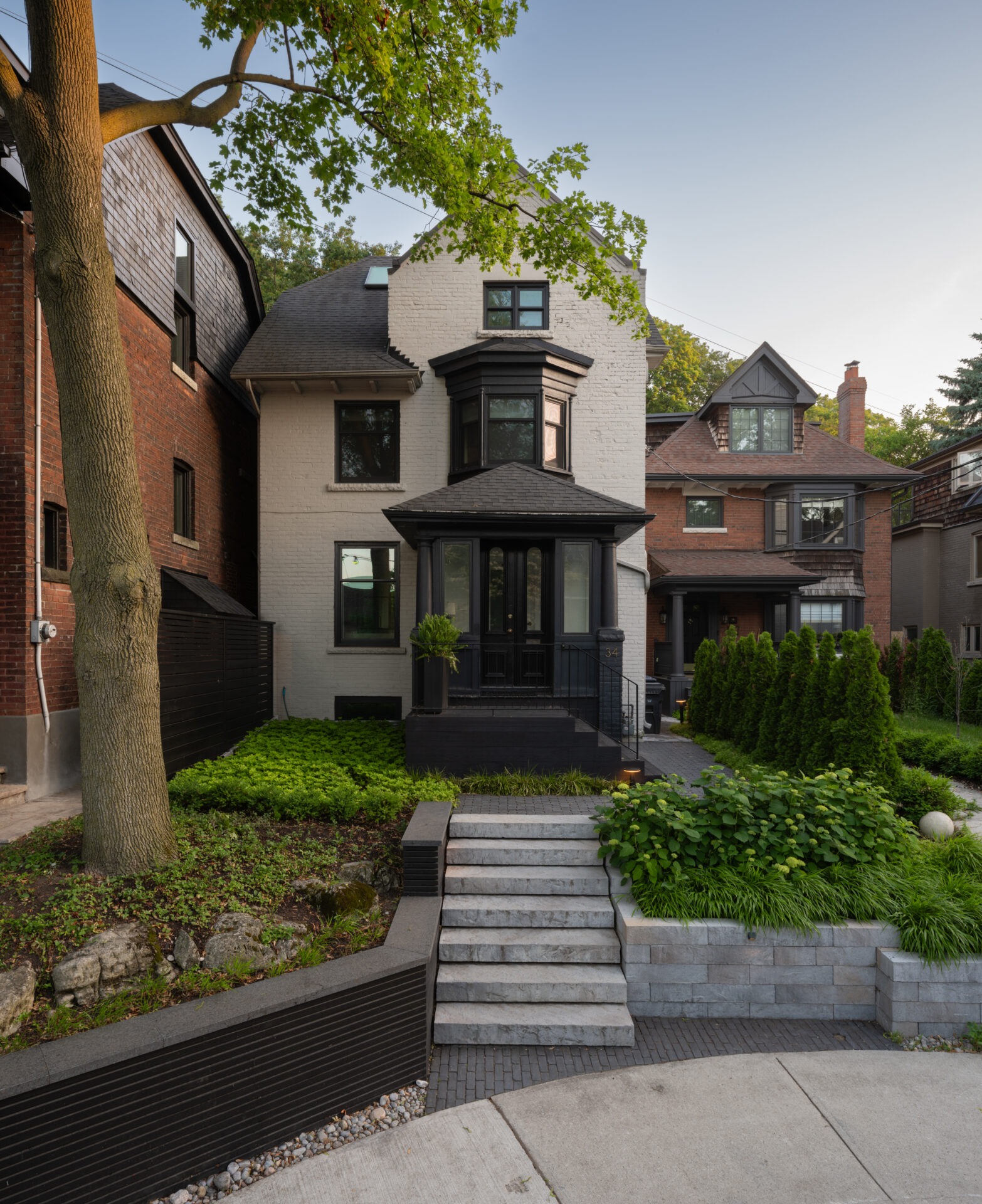 A charming two-story brick house with a lush garden and stone stairs, surrounded by greenery and neighboring homes in a residential area.
