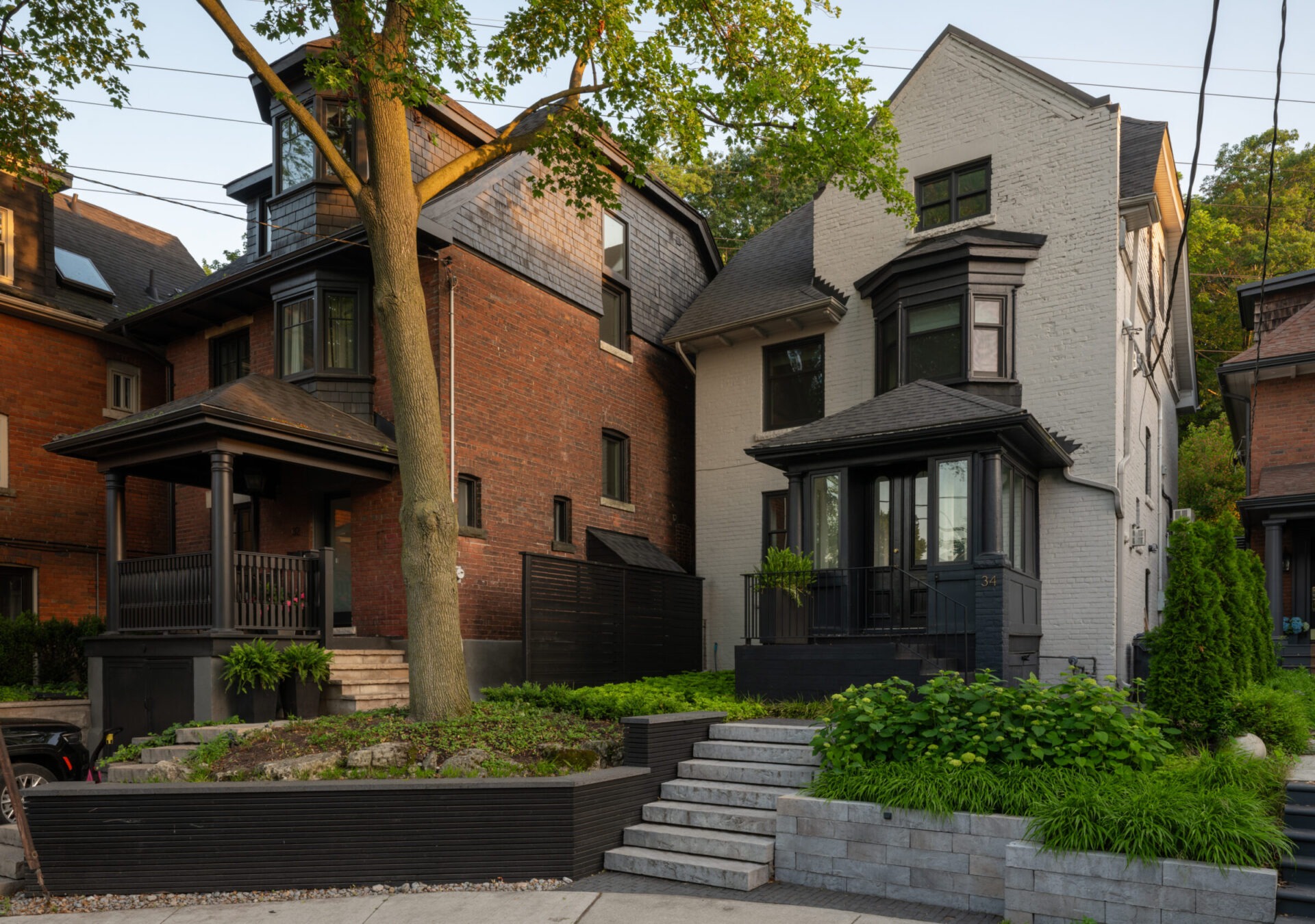 Two adjacent, charming brick houses with porches, surrounded by lush greenery and tree, under a clear sky, creating a peaceful urban neighborhood scene.