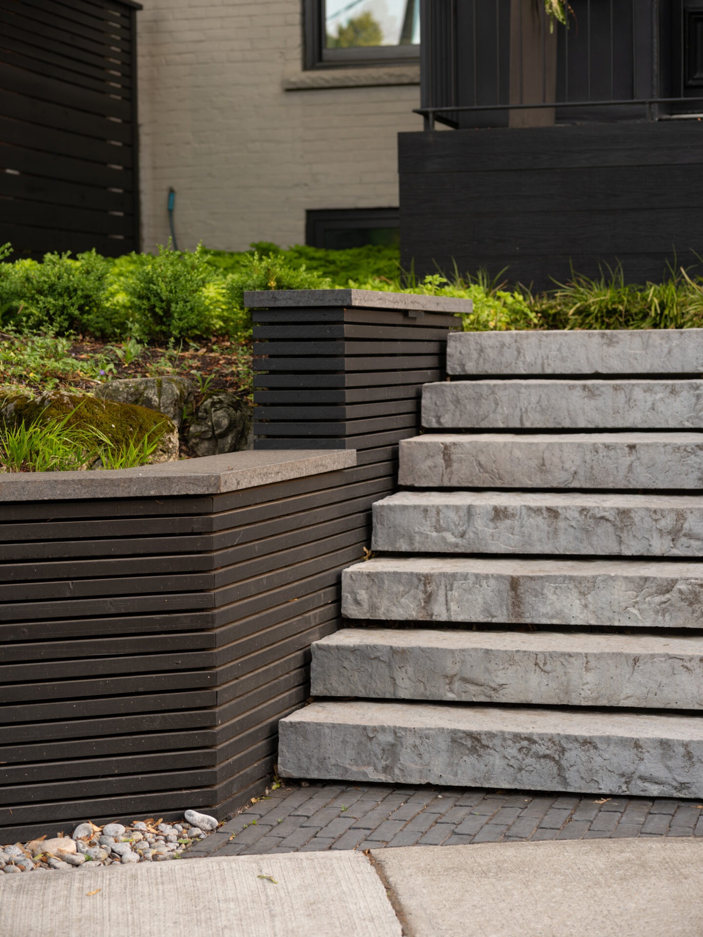 Gray stone steps lead up to a modern building with lush greenery, contrasting against black wooden panels and a beige brick wall.