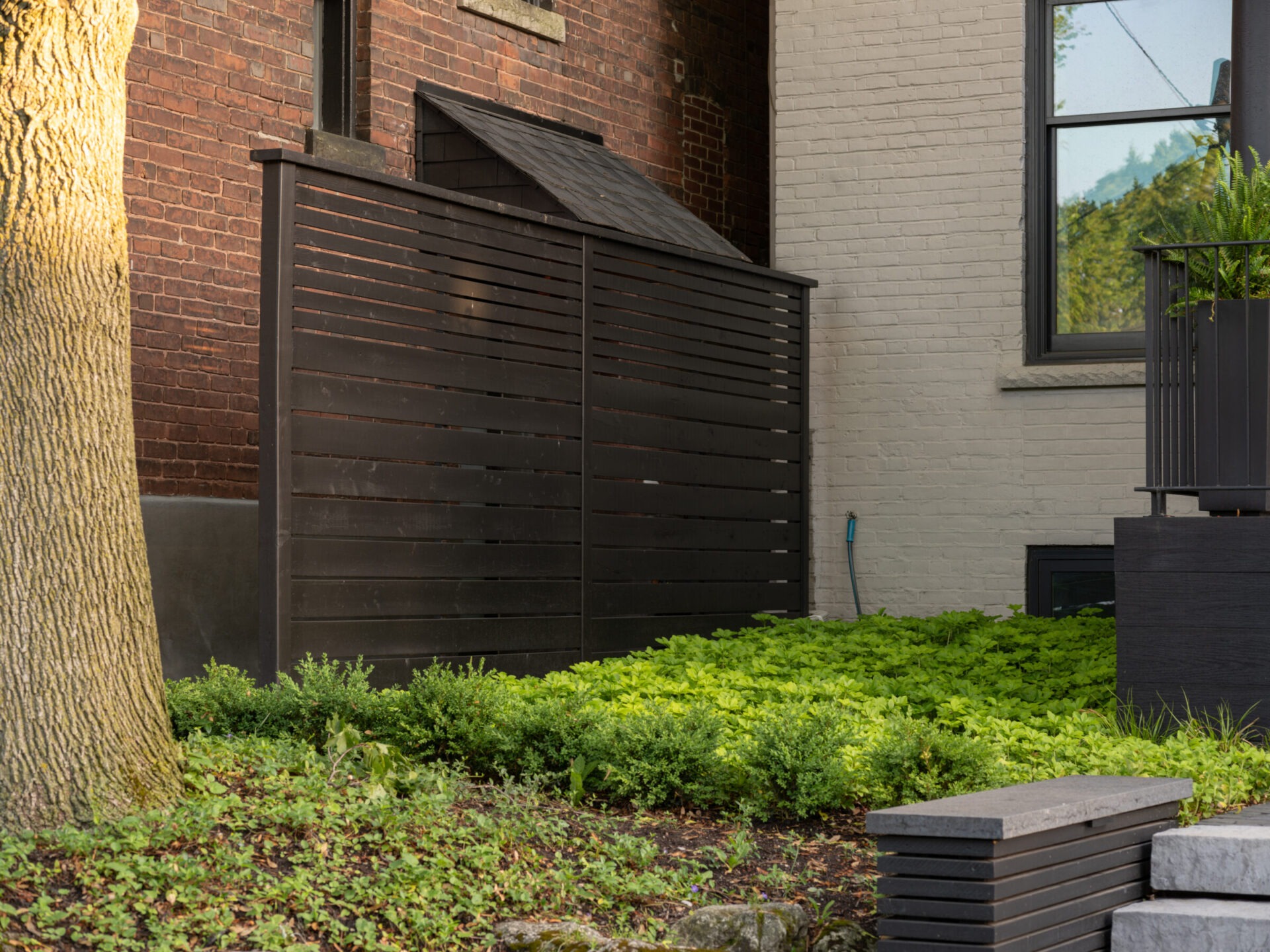 A modern, wooden privacy fence is set against a brick wall and lush garden, with a large tree in the foreground.