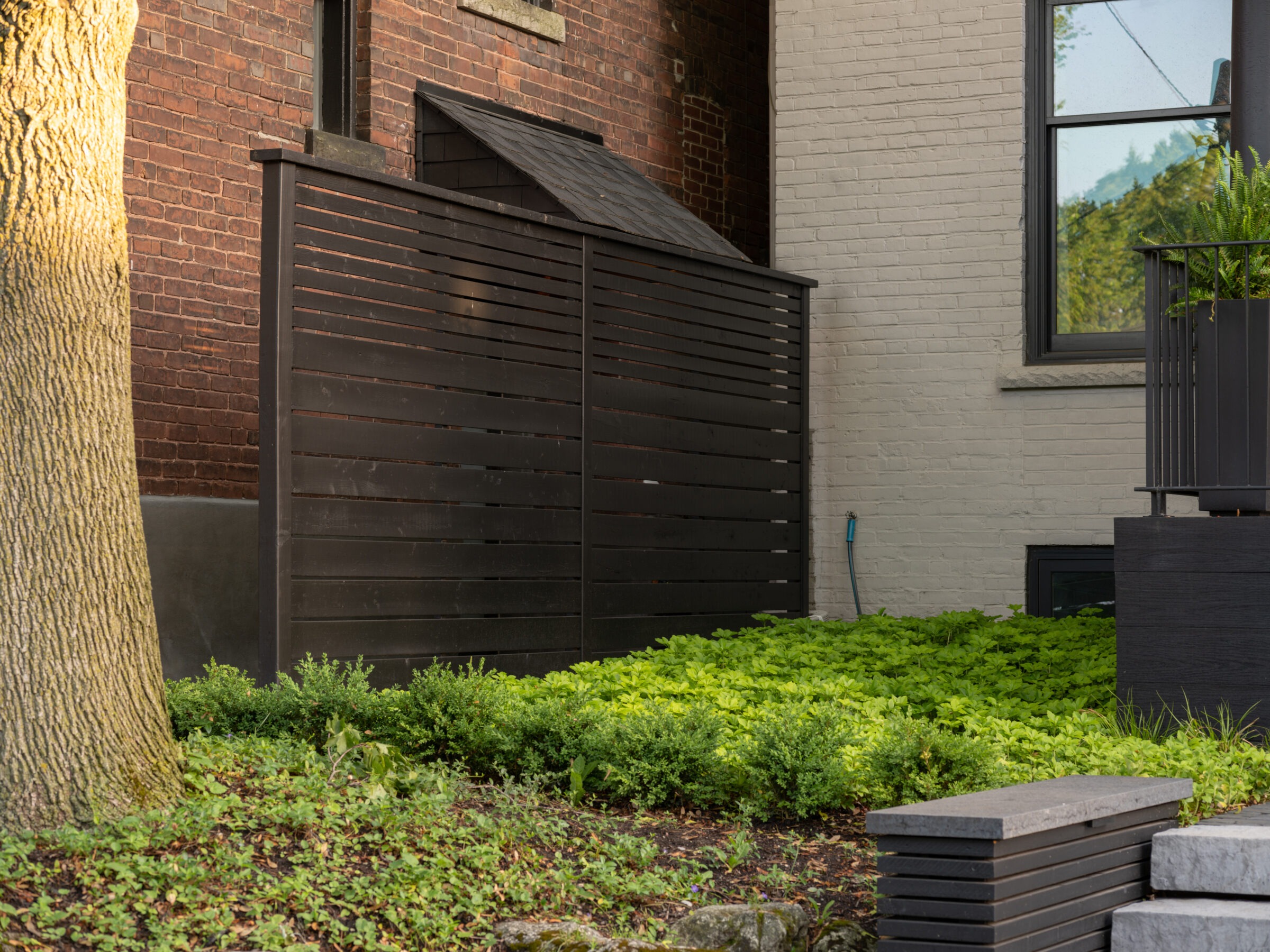 A modern, wooden privacy fence is set against a brick wall and lush garden, with a large tree in the foreground.
