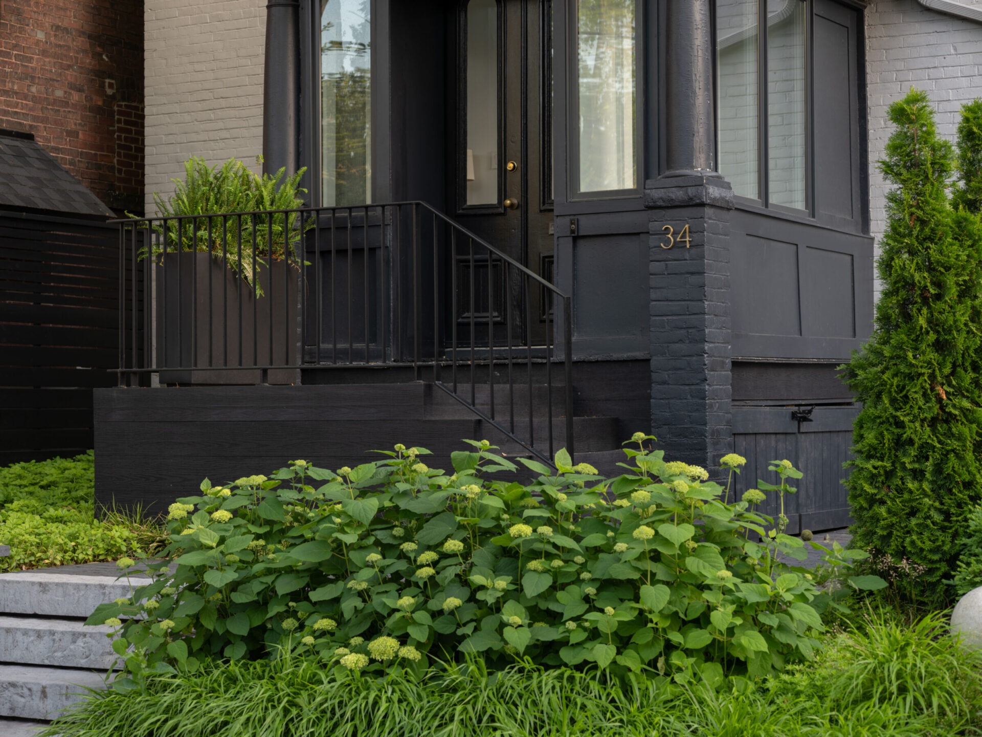 A modern black-painted entrance with steps, metal railing, and lush green plants surrounds the setting. The house number 