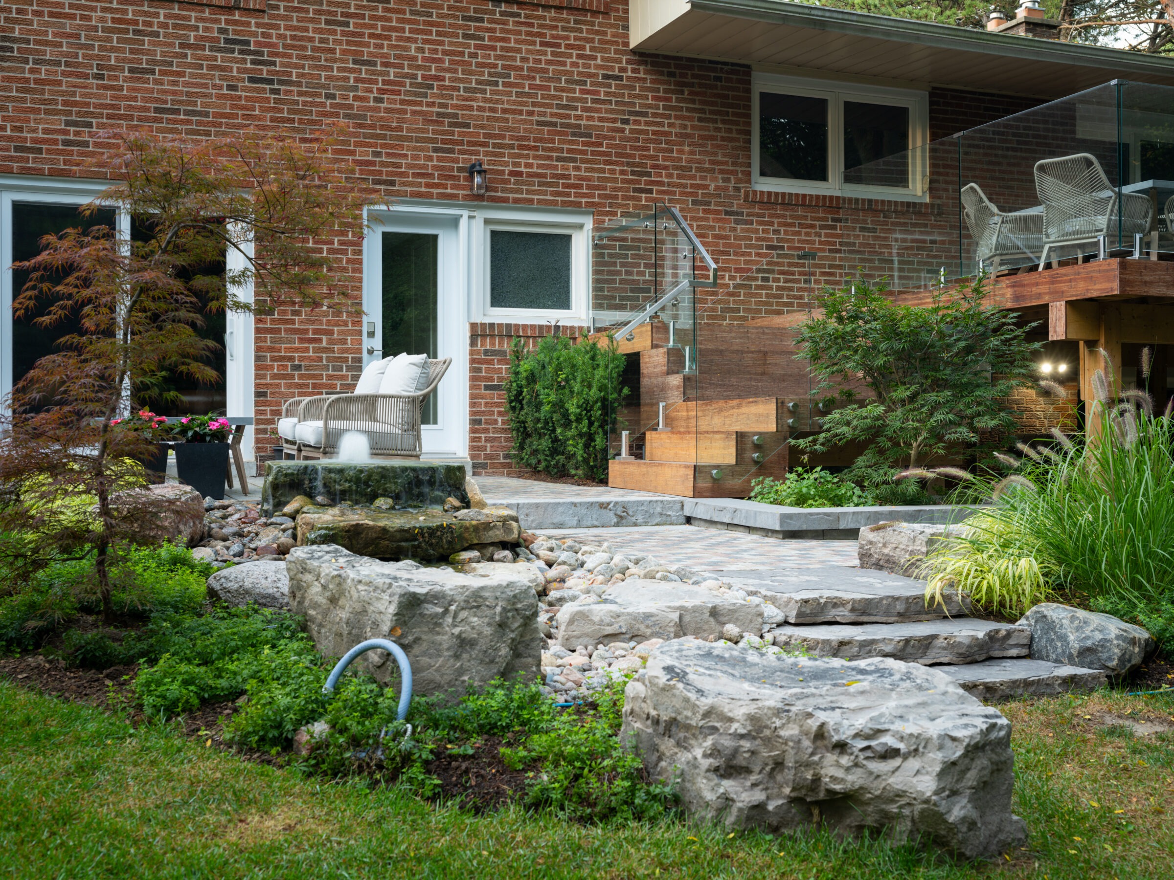 A modern brick house features a landscaped garden with large rocks, greenery, and a stylish seating area on a raised patio.