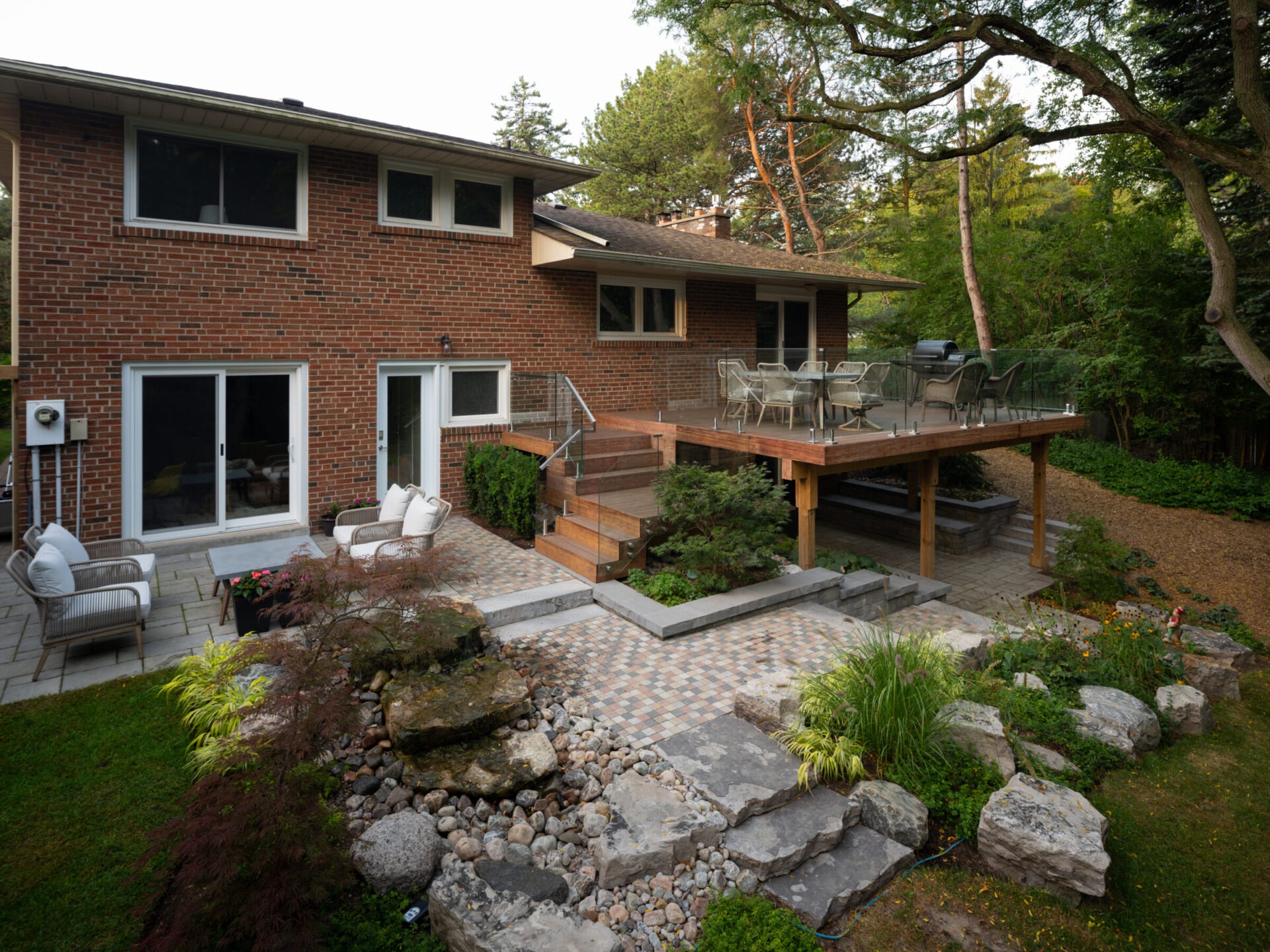 A brick house with a raised wooden deck and patio, surrounded by lush greenery and decorative stone landscaping in a peaceful setting.
