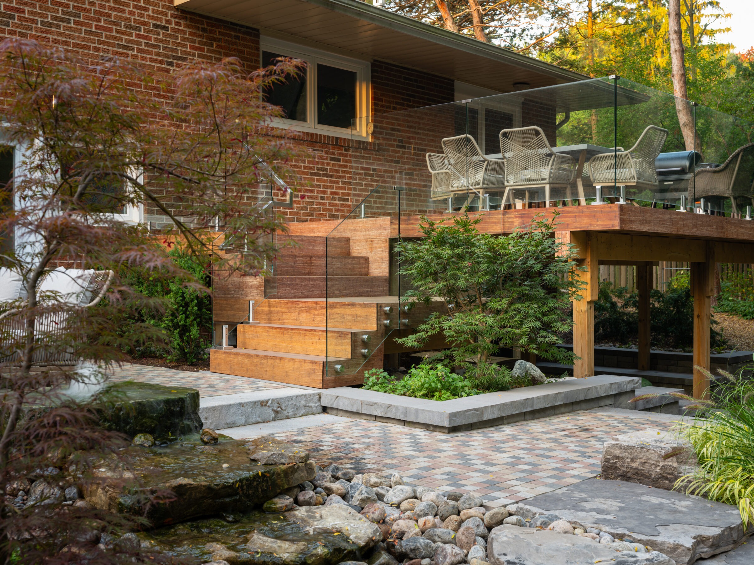 A modern backyard features a wooden deck with glass railing, patio furniture, lush greenery, stone pathway, and a small rock water feature.