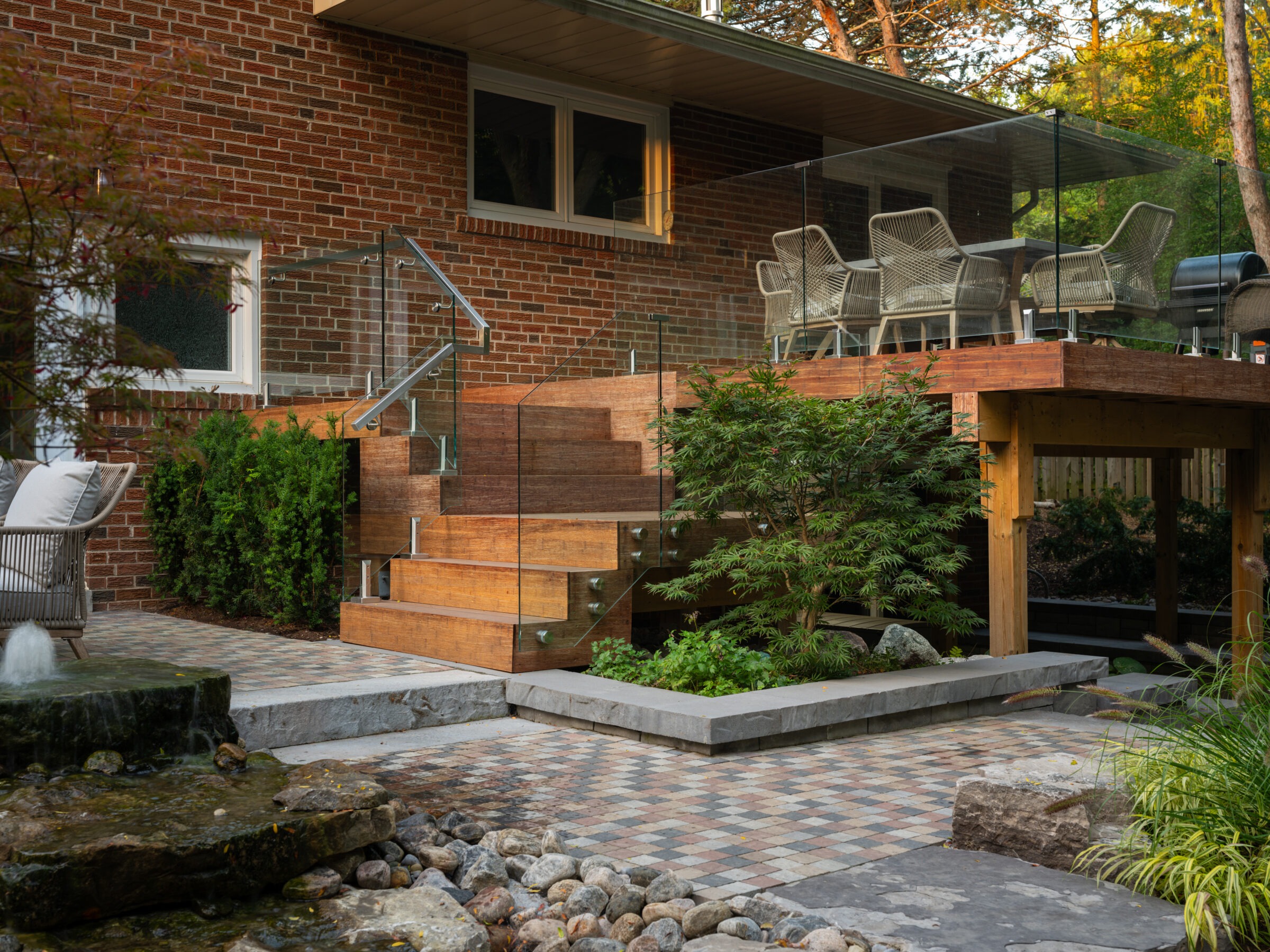 Modern backyard with wooden deck, glass railings, and seating area. Brick house backdrop, landscaped garden, water feature, and stone pathway enhance tranquility.