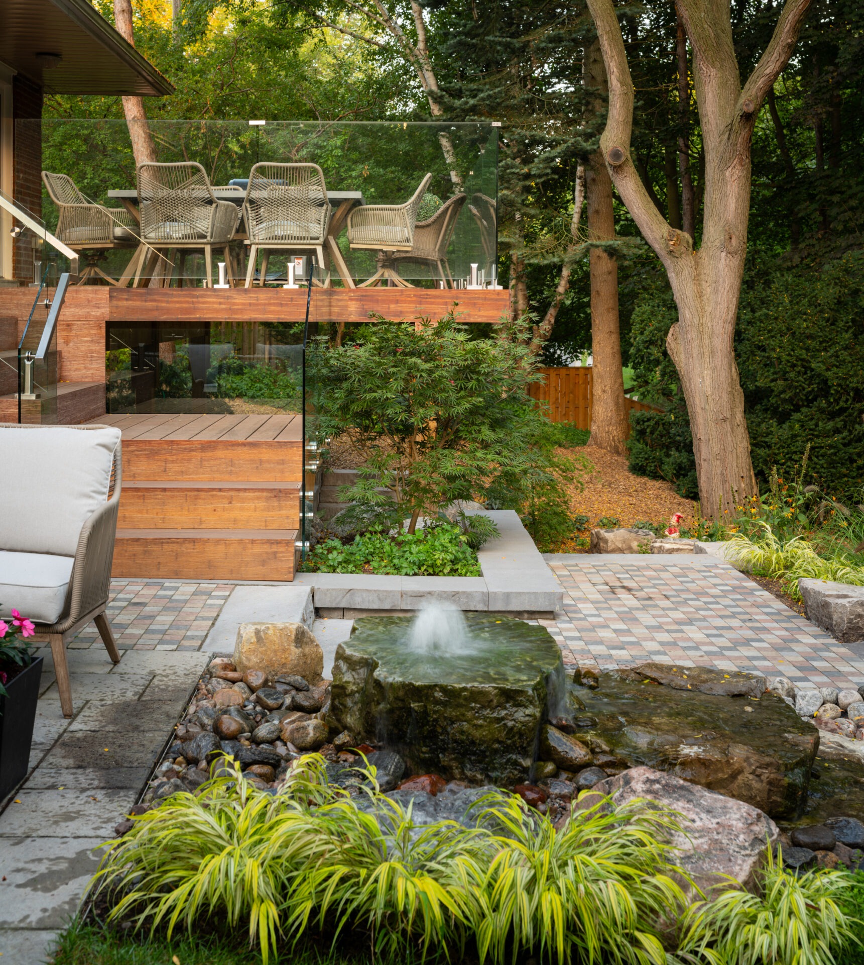 A serene backyard with patio furniture, lush greenery, and a small water feature fountain, surrounded by trees and landscaped stone pathways.