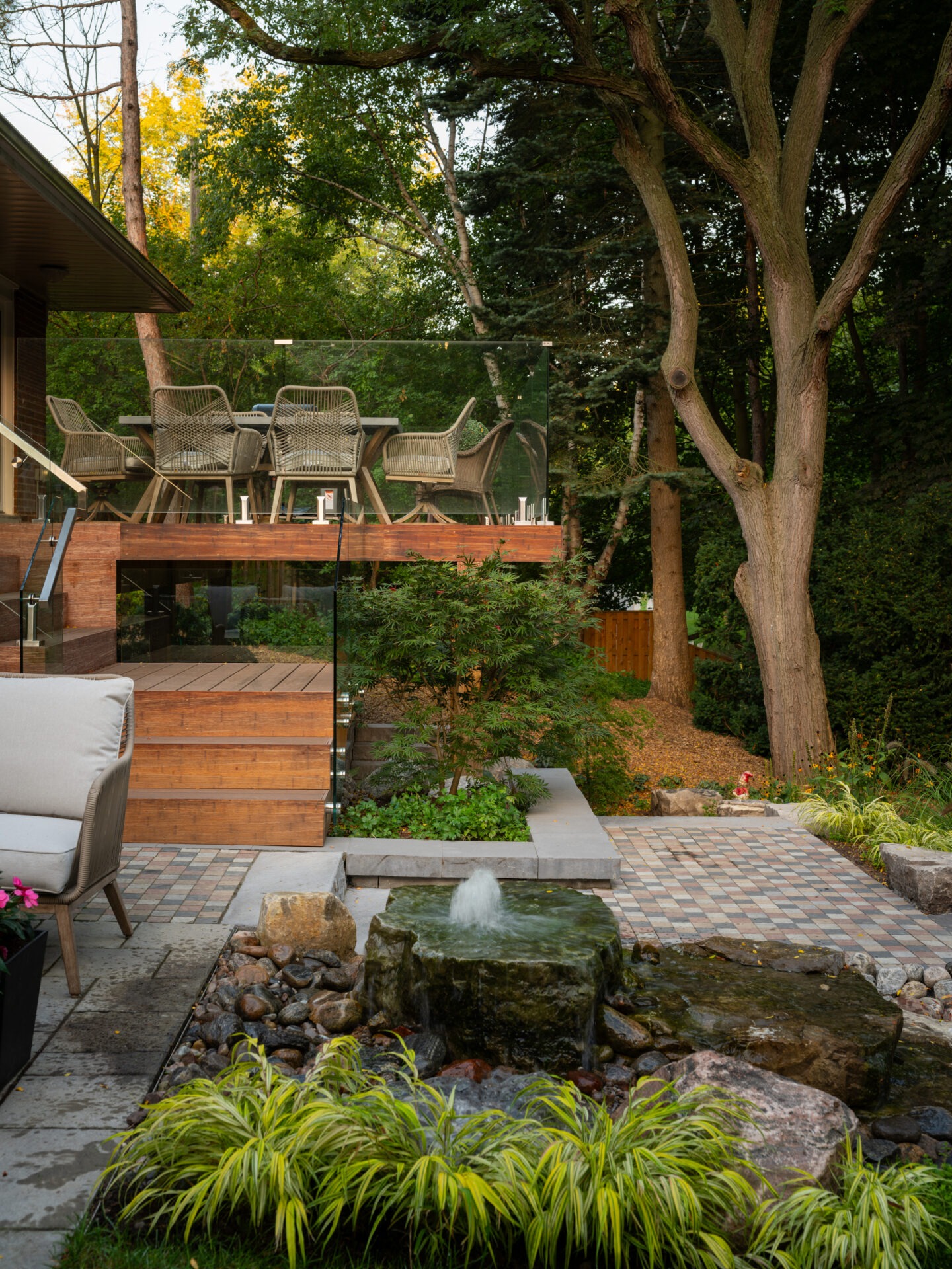 Modern backyard with patio furniture, a glass railing, and a small fountain. Surrounded by lush greenery and tall trees in tranquil setting.