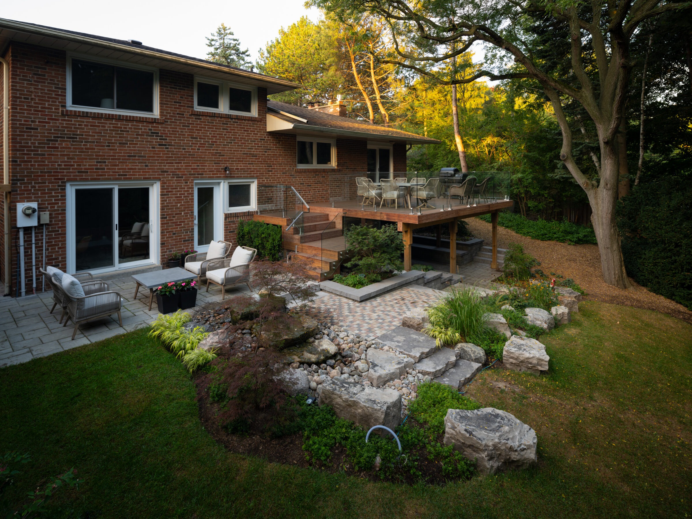 A modern brick house with a spacious patio, comfortable seating, landscaped garden, and large trees in a serene, sunlit setting.