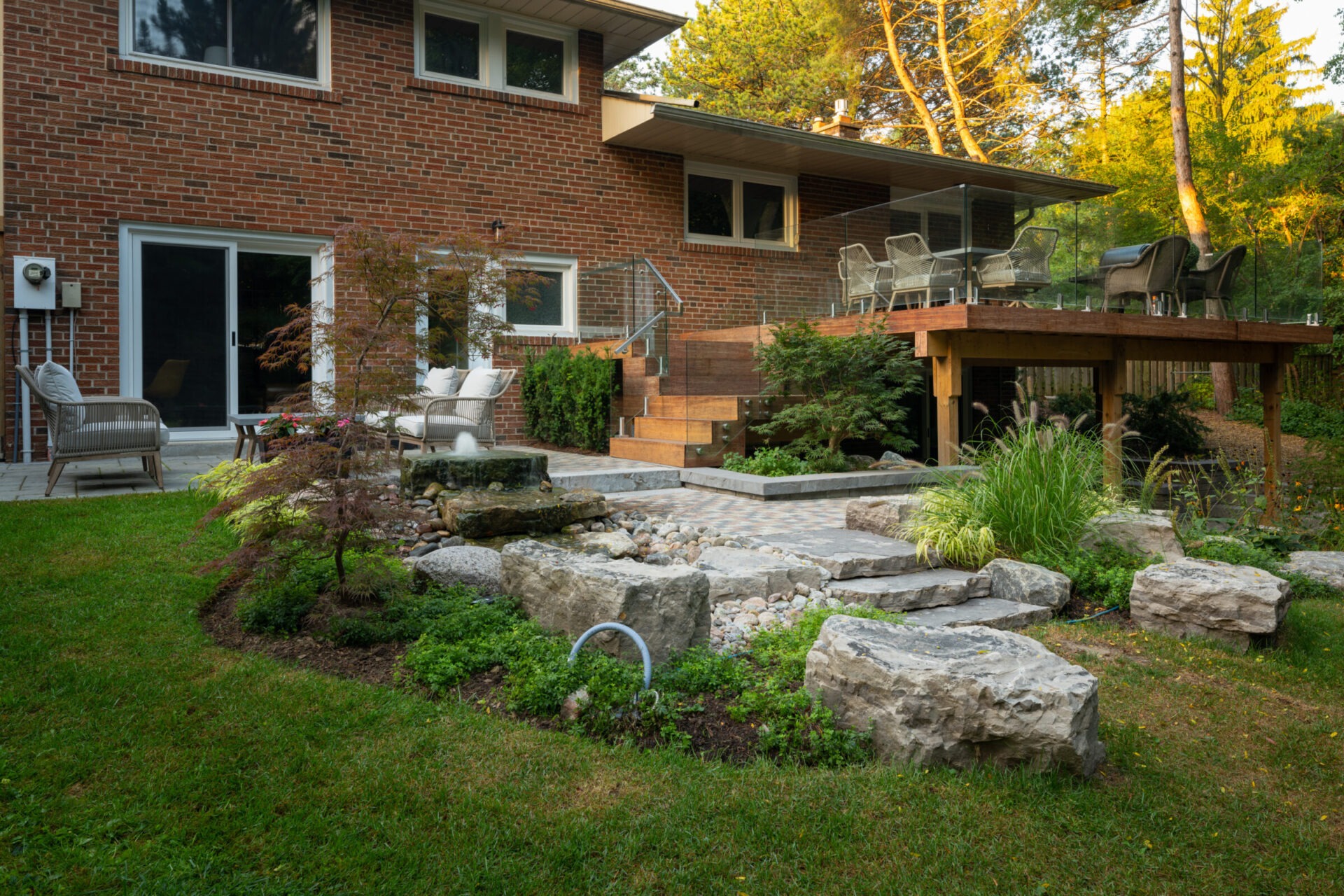 Brick house with a landscaped backyard featuring a stone path, trees, and outdoor seating area under a wooden deck surrounded by greenery.