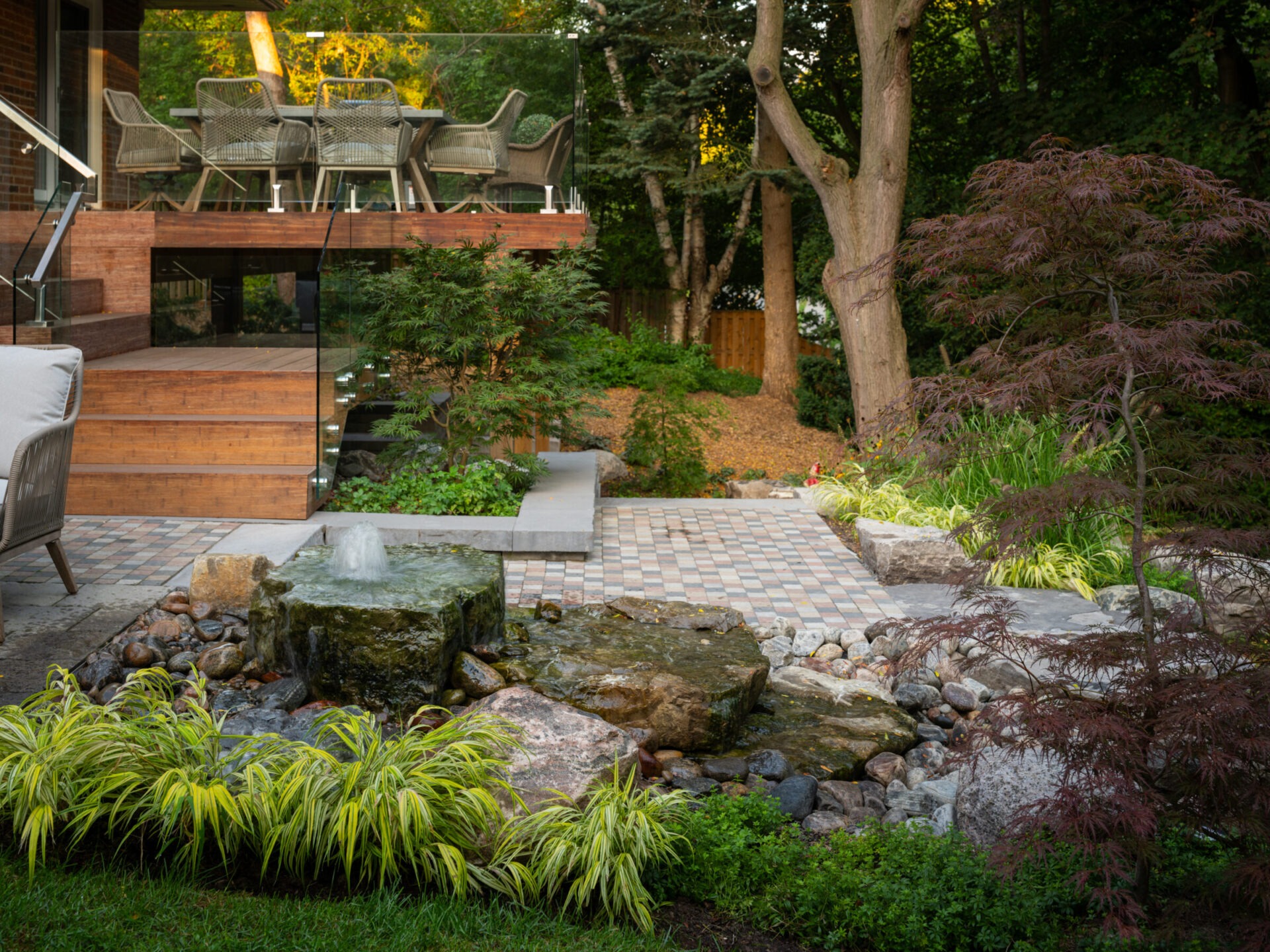 Lush garden with a stone fountain, wooden deck, glass railing, and outdoor dining set surrounded by trees and foliage, creating a serene atmosphere.