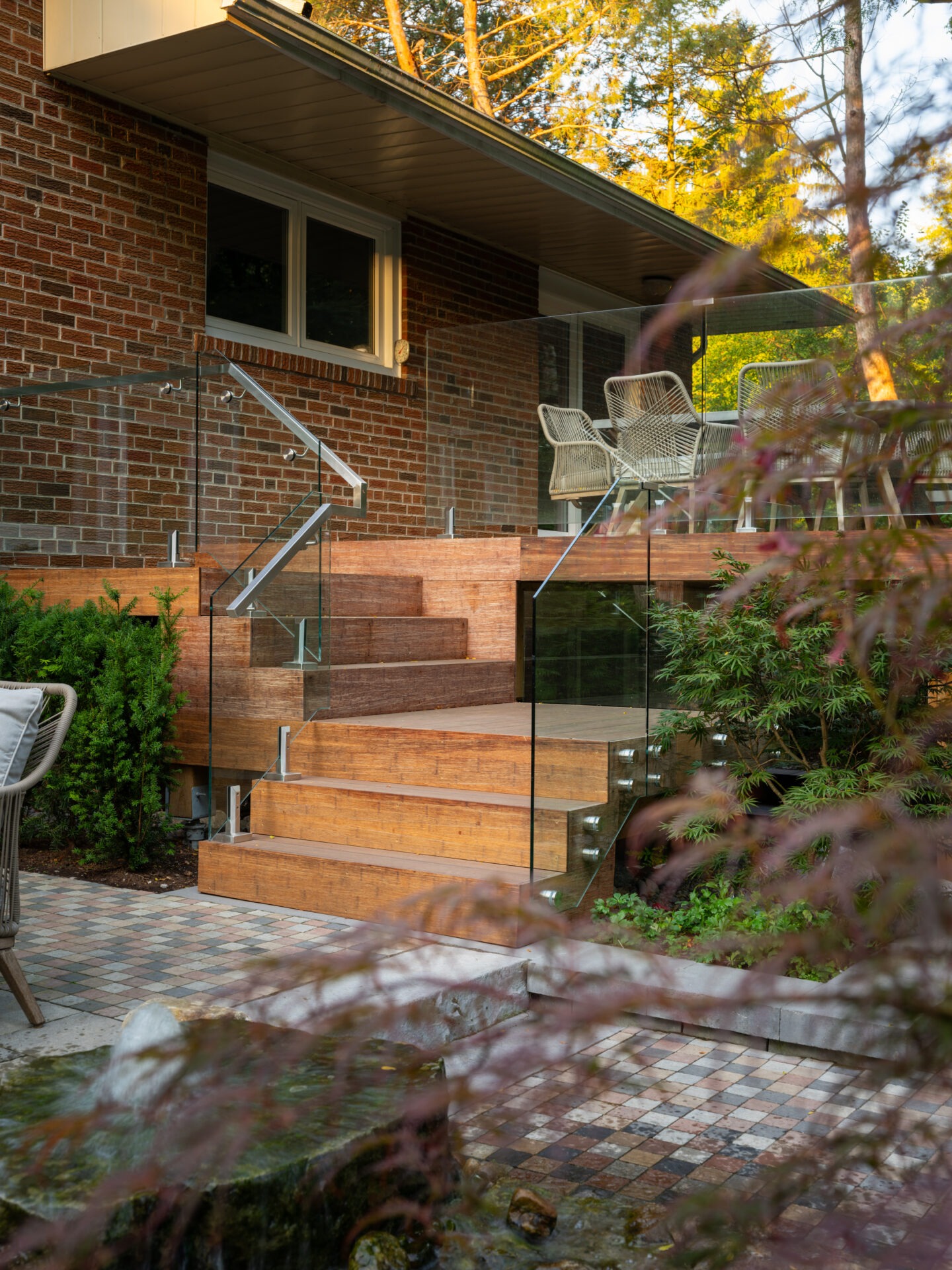 Modern patio with wooden steps, glass railing, and wicker chairs. Surrounded by greenery and a water feature, creating a tranquil outdoor space.