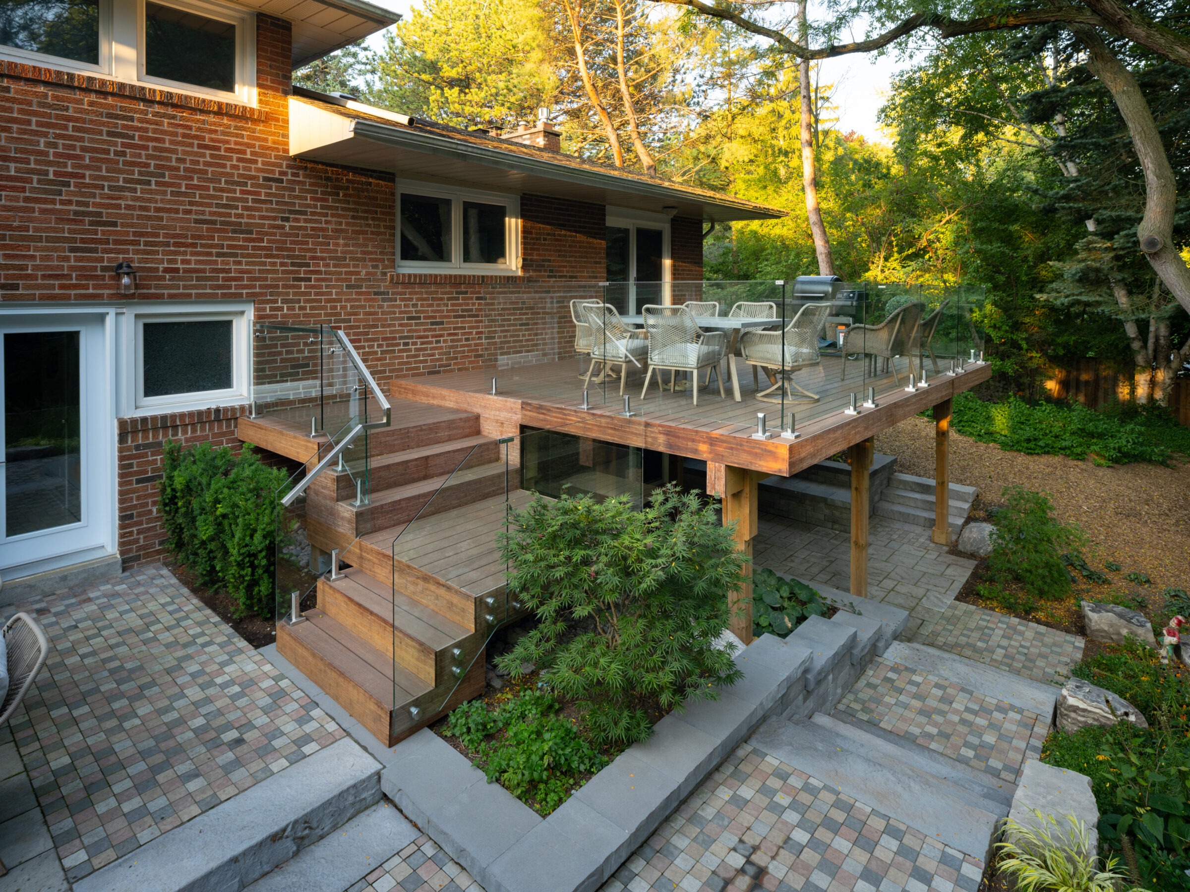 Brick house with a wooden patio and glass railing, featuring outdoor seating. Surrounded by lush greenery and stone pathways.