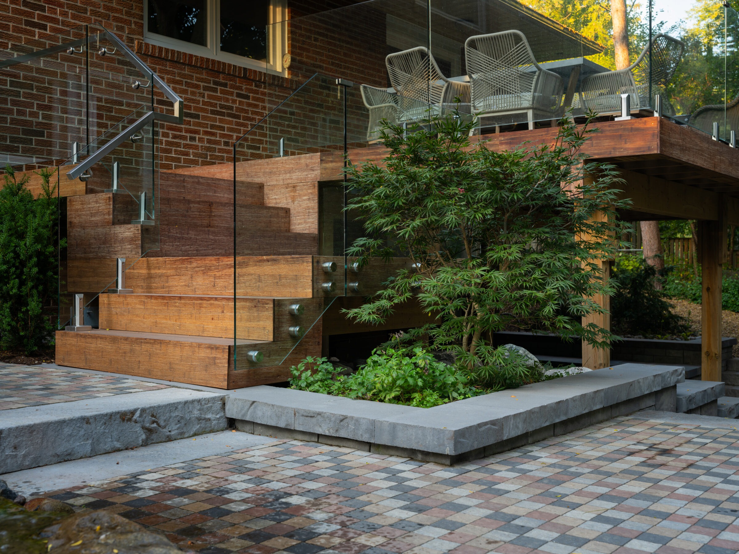 Modern terrace with wooden steps and glass railing, surrounded by greenery. Tiled patio features geometric patterns, enhanced by ambient natural light, creating a serene outdoor atmosphere.
