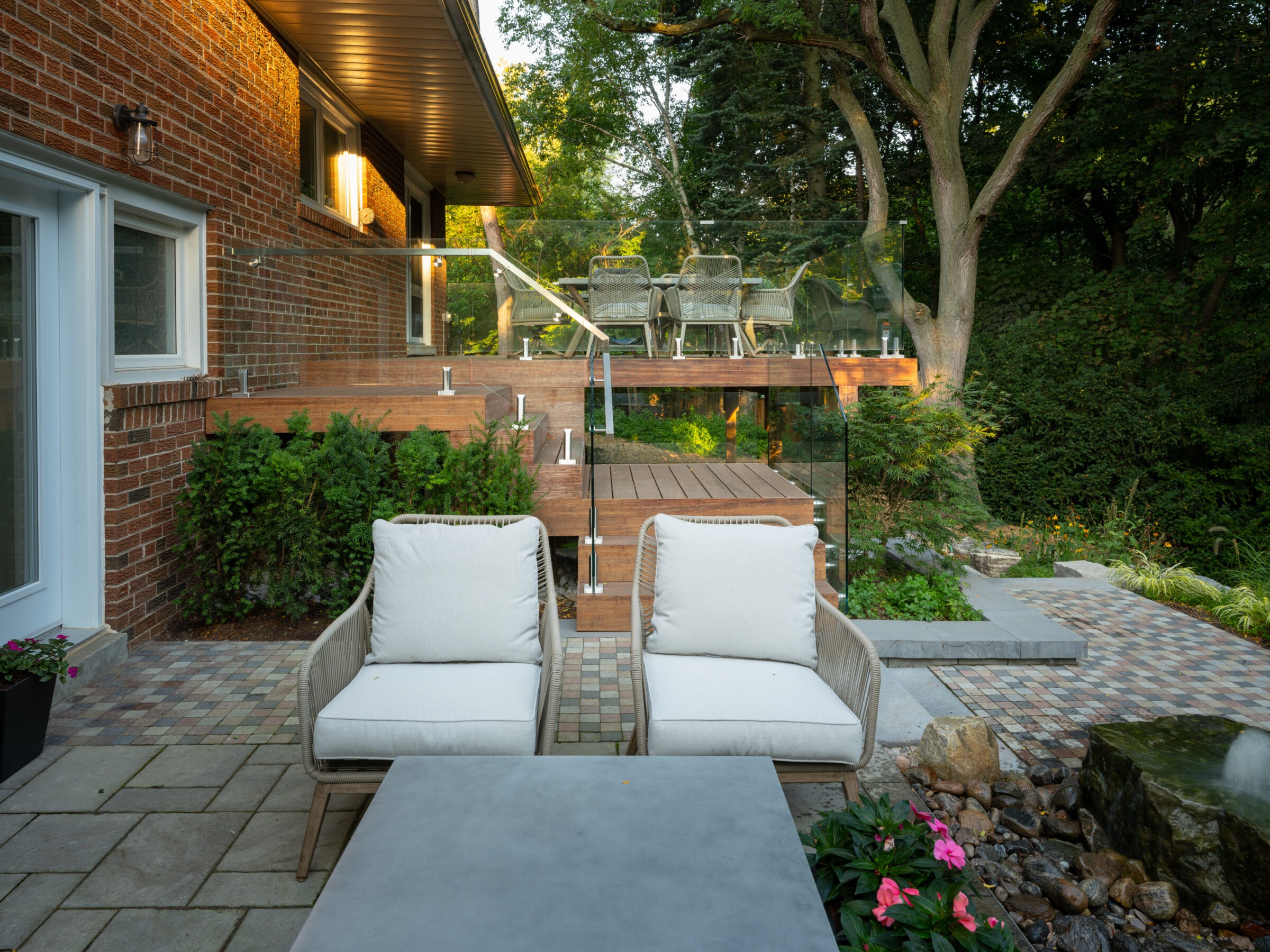 A cozy outdoor patio with cushioned chairs, brick and wood elements, surrounded by lush greenery and a small garden area.