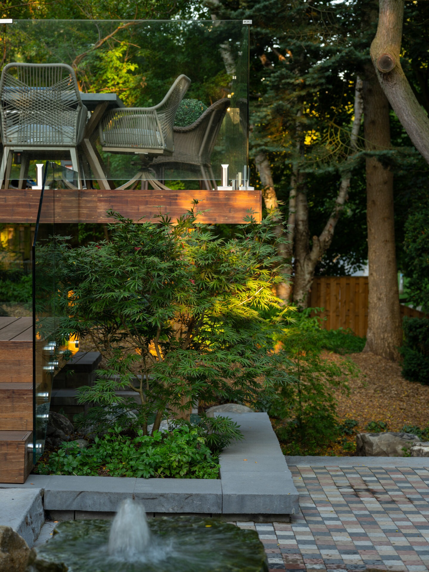 A modern outdoor patio with wicker furniture overlooks a landscaped garden, featuring a small fountain and lush greenery under warm evening light.