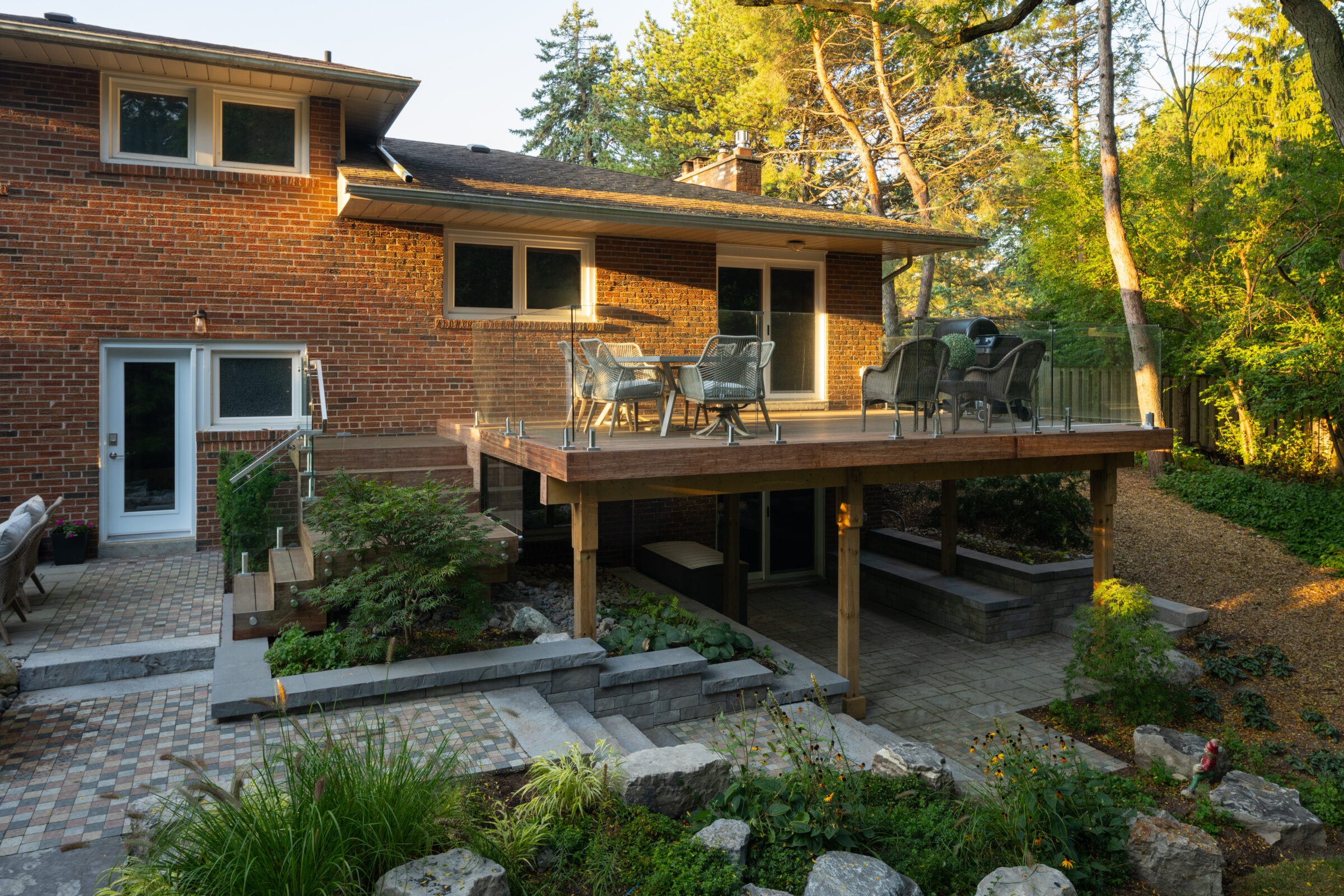 Brick house with elevated wooden deck, glass railings, and outdoor seating. Surrounded by trees and gardens, creating a serene, natural atmosphere.