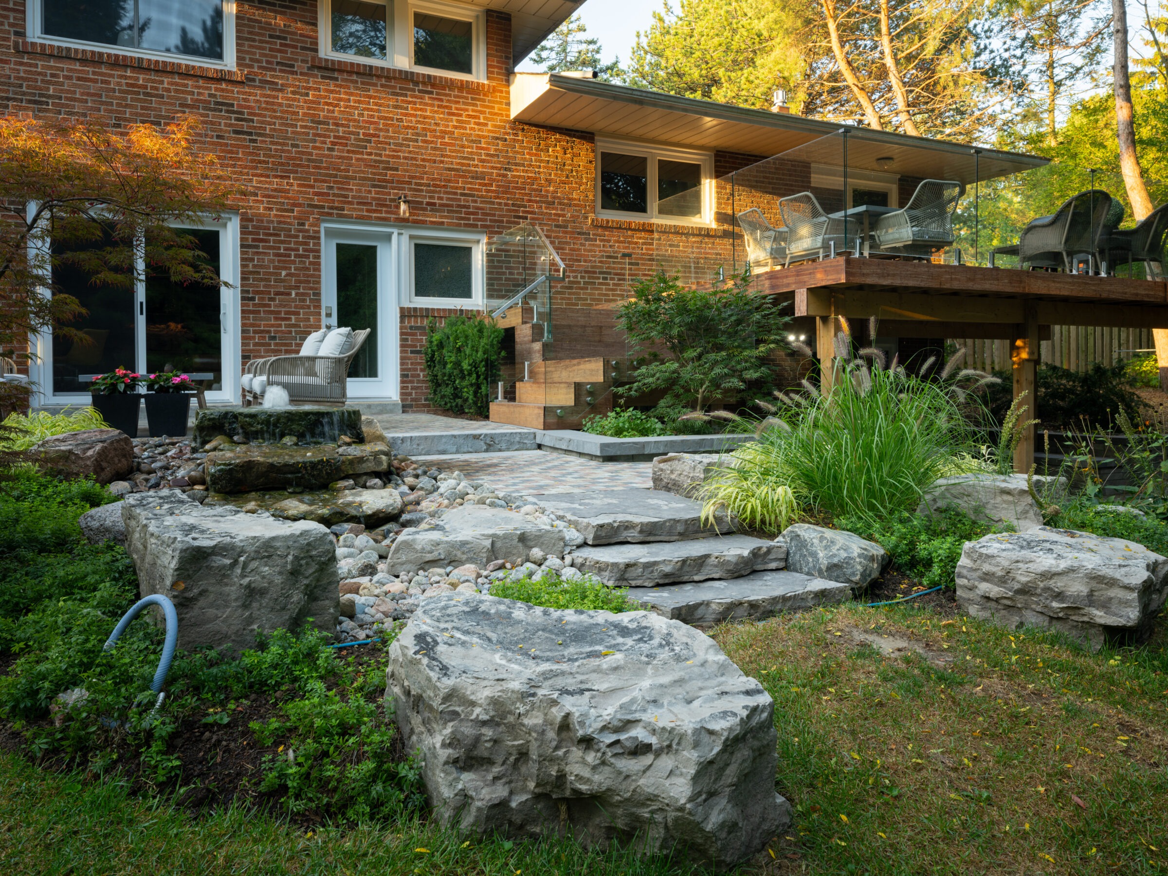 A modern brick house with a wooden deck, outdoor furniture, and landscaped garden featuring stone steps, large rocks, and lush greenery.