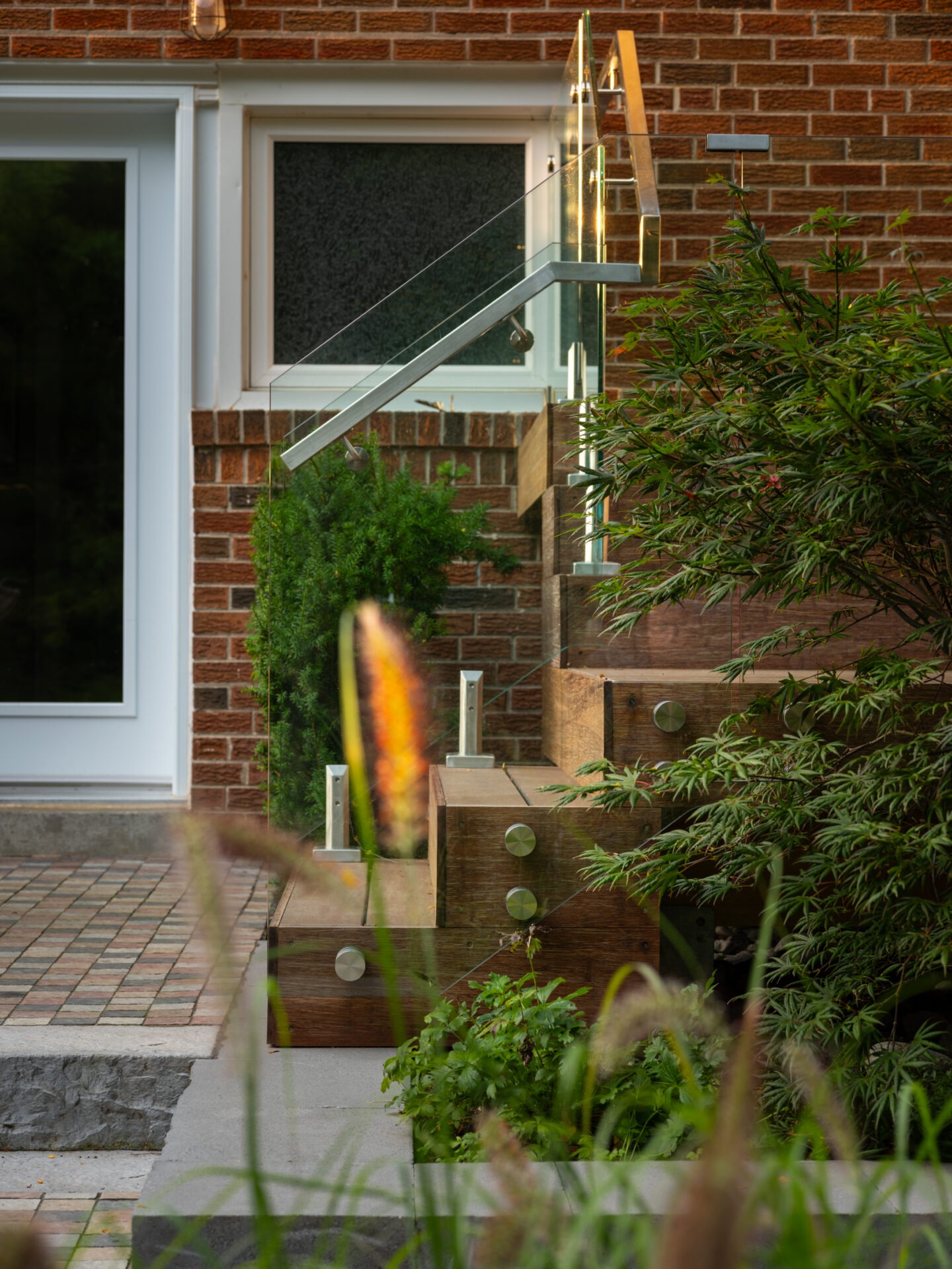 Brick house entrance with wooden steps, glass railing, and lush greenery. Modern design integrates natural elements with sleek architectural features for a welcoming ambiance.