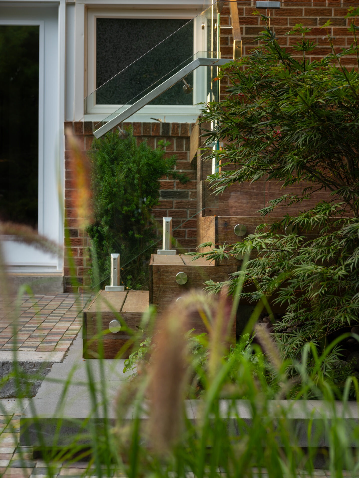 Modern architectural entrance with glass railing, wooden accents, and lush greenery. Clean lines, brick wall, and pavers create a stylish, welcoming ambiance.