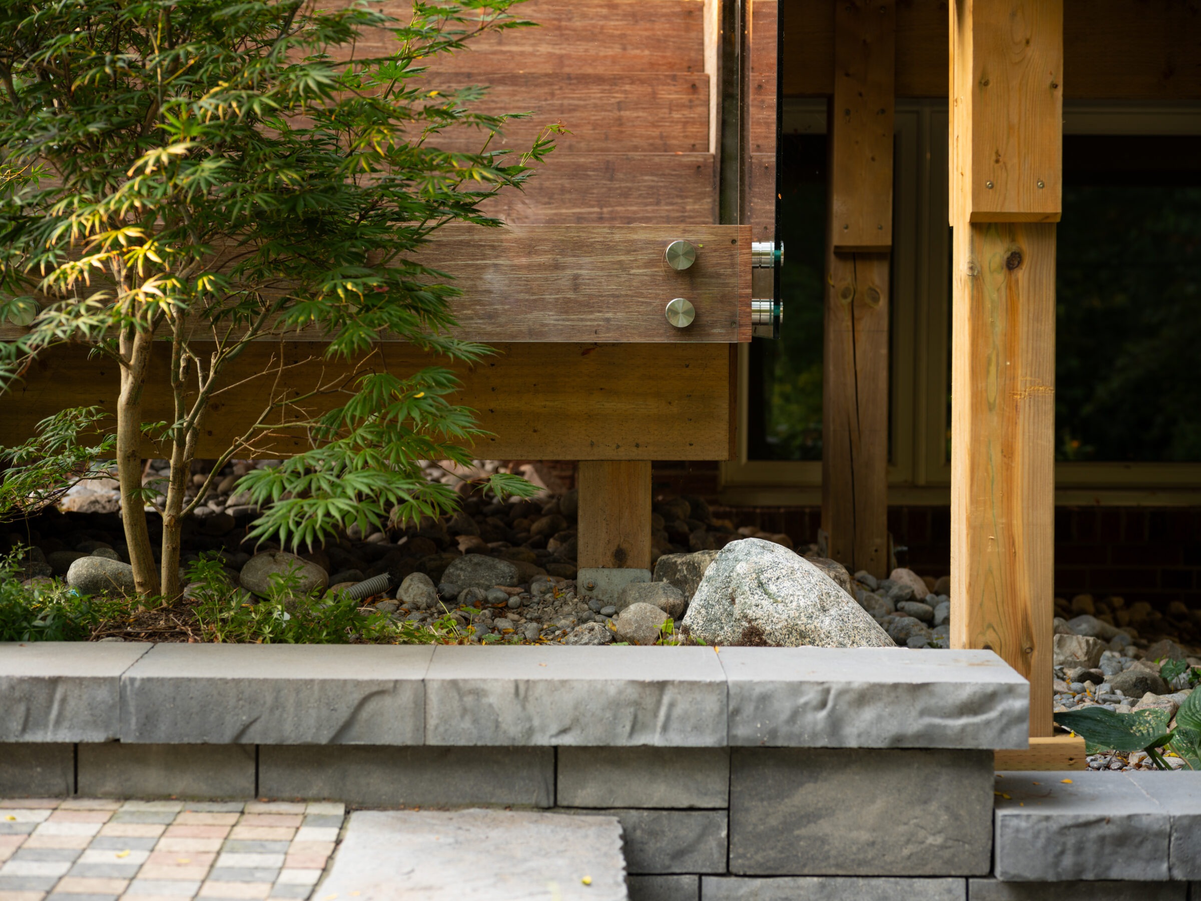 A wooden structure with metal accents, surrounded by stones and lush greenery, sits on a stone-paved area, creating a serene, natural setting.