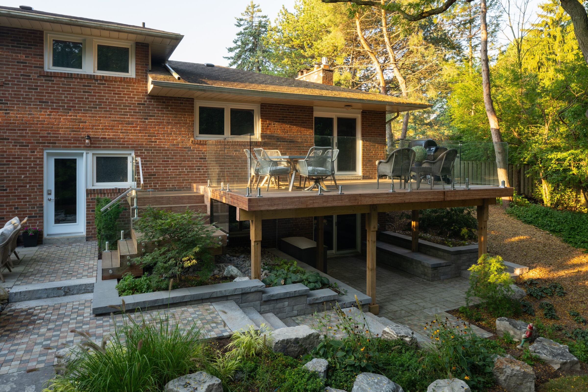 Brick house with elevated deck, surrounded by lush greenery. Outdoor seating area with chairs. Stone pathway and landscaped garden enhance the serene setting.