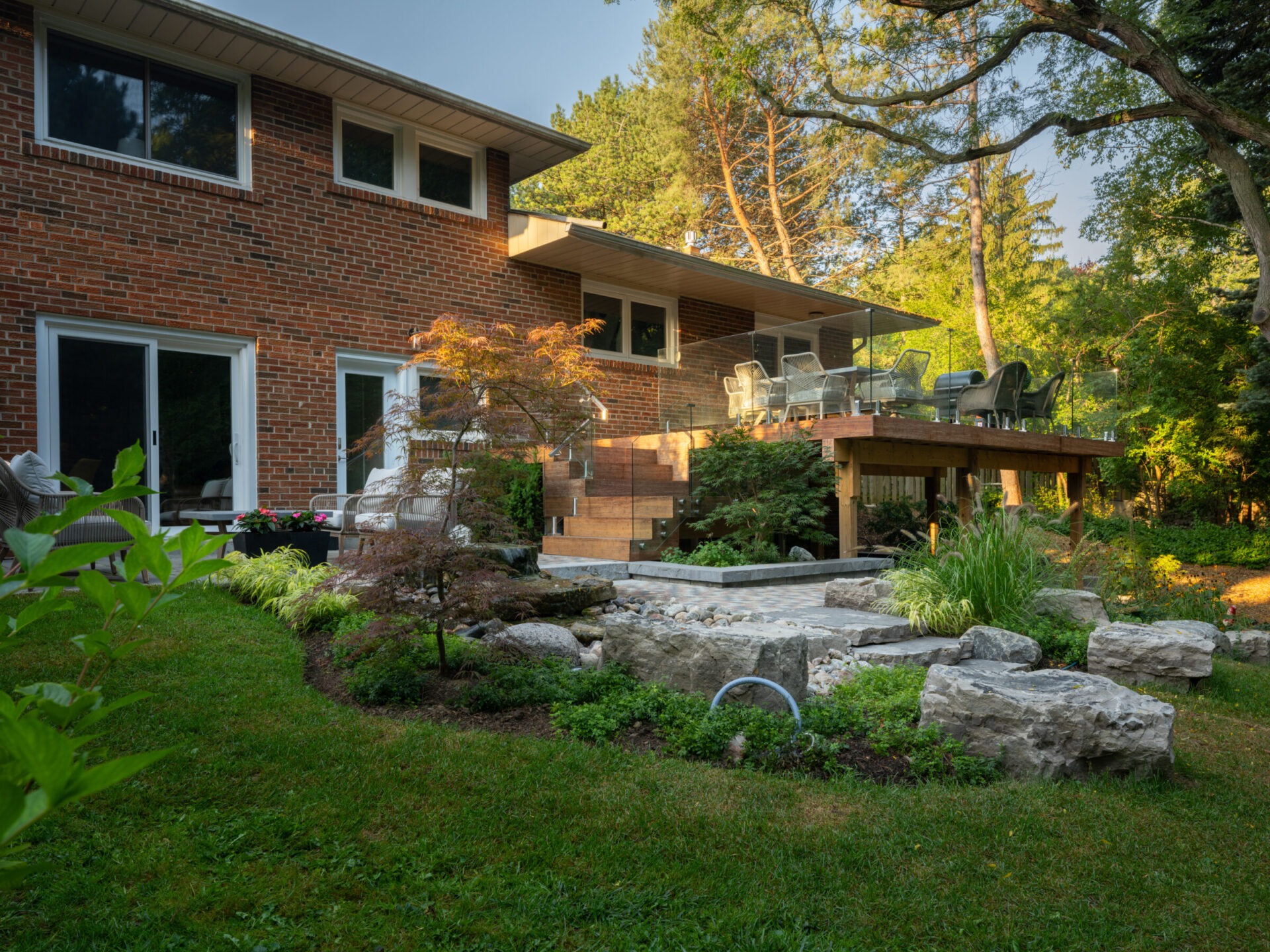 A two-story brick house with a wooden deck overlooks a landscaped garden with rocks and lush greenery, surrounded by tall trees.