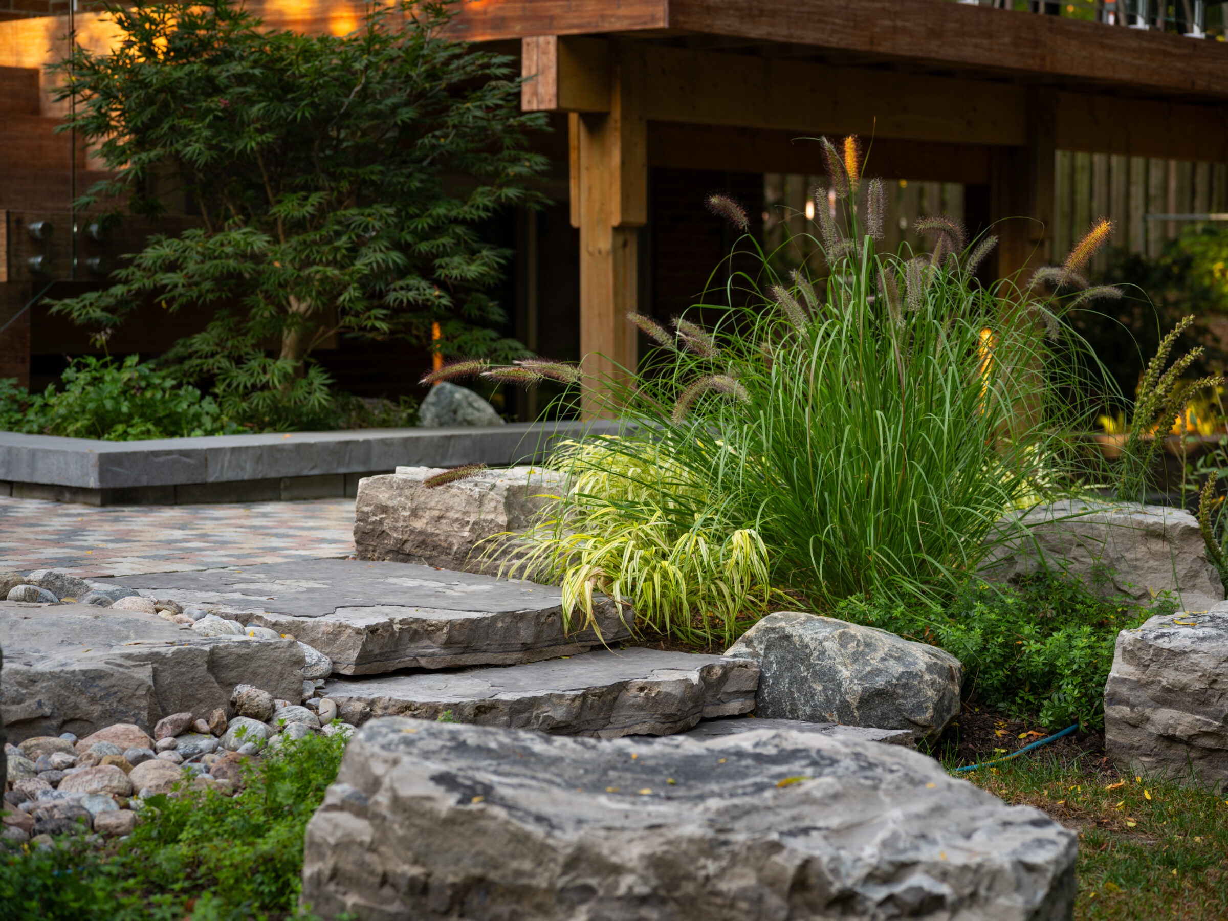 A tranquil garden scene with rocks, lush grasses, and a wooden structure featuring modern landscaping and natural elements.