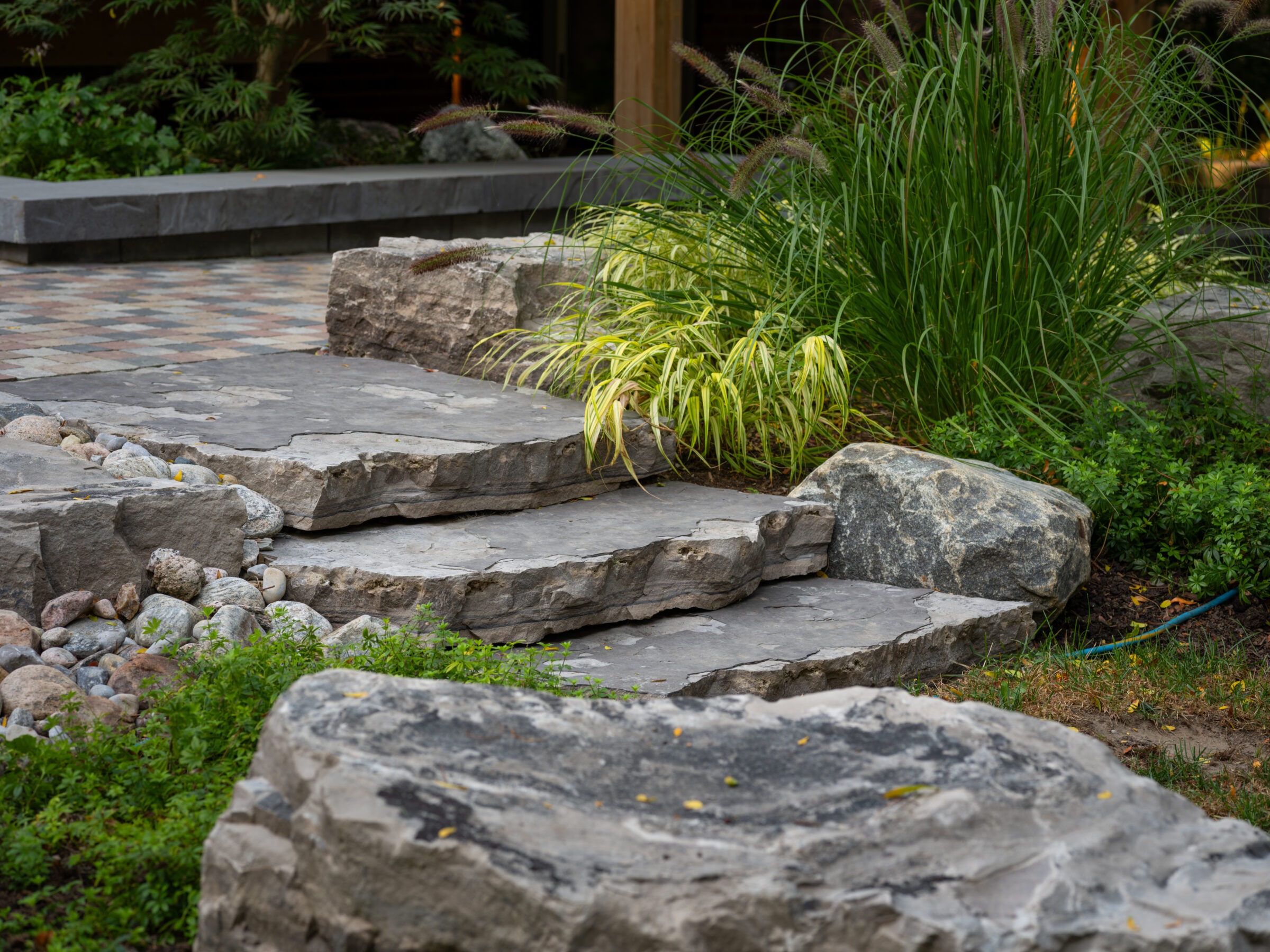 A serene garden landscape featuring large stone steps, lush green plants, and a patterned stone pathway amidst natural rock formations.