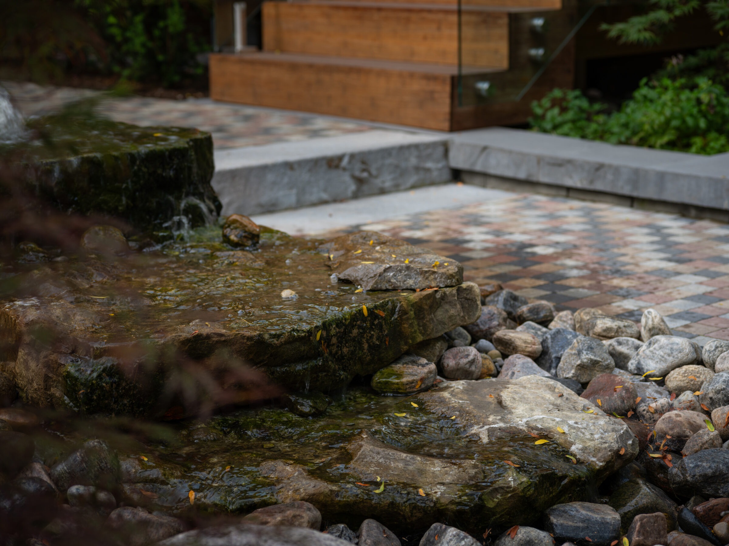 A serene garden scene with a small rock waterfall, surrounded by pebbles and greenery, next to a wooden staircase and checkered pathway.